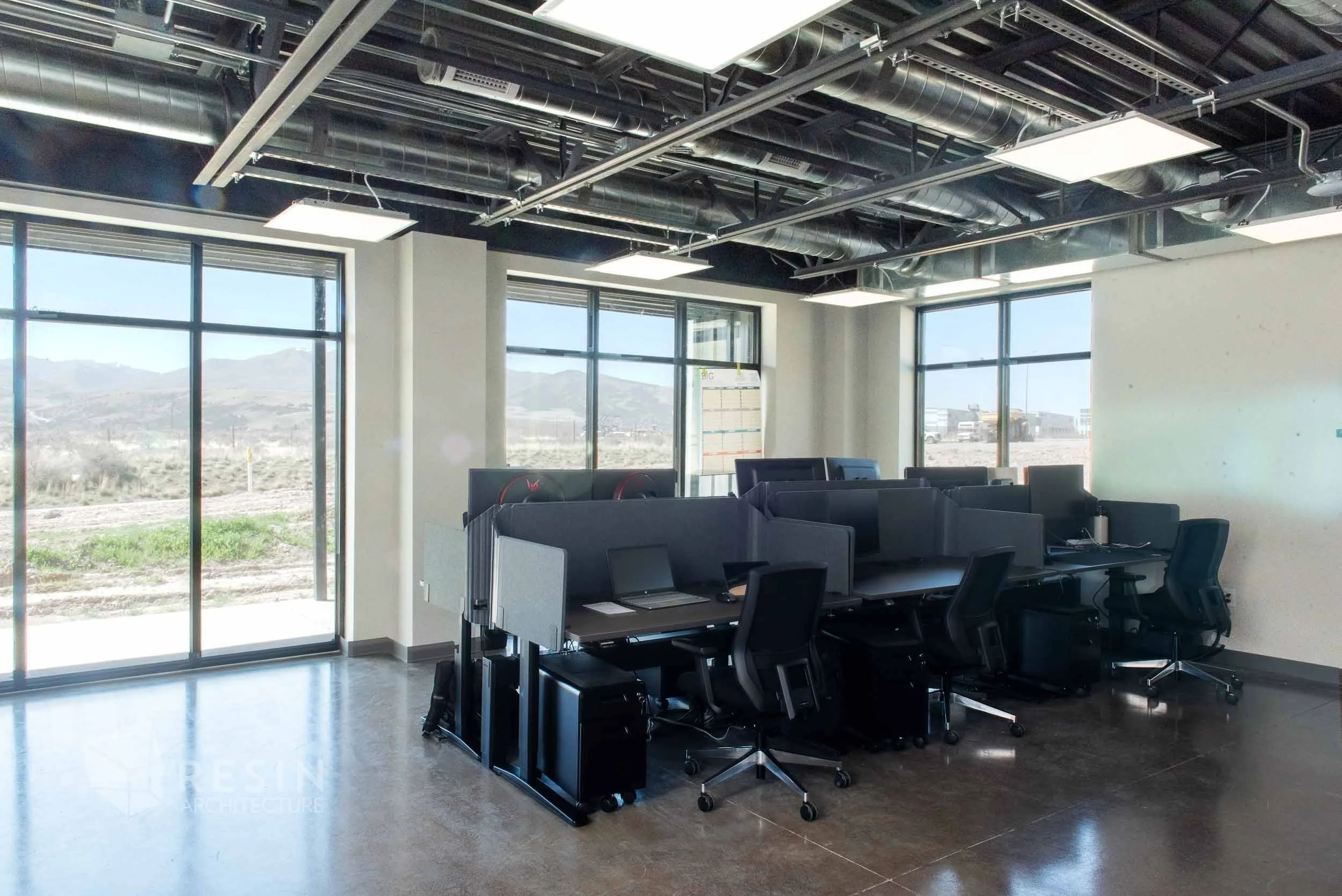 Modern office with large windows, black desks with monitors, and ergonomic chairs, with an open ceiling design showing ventilation ducts and lighting fixtures.