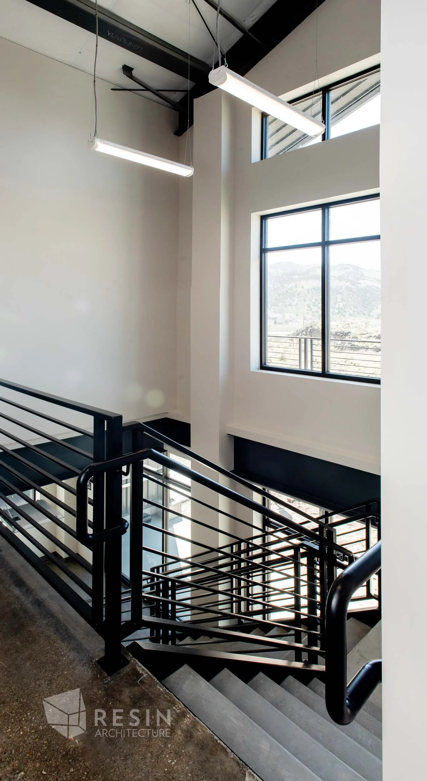 Interior view of a modern stairwell with black metal railings, white walls, large windows, and overhead fluorescent lights.