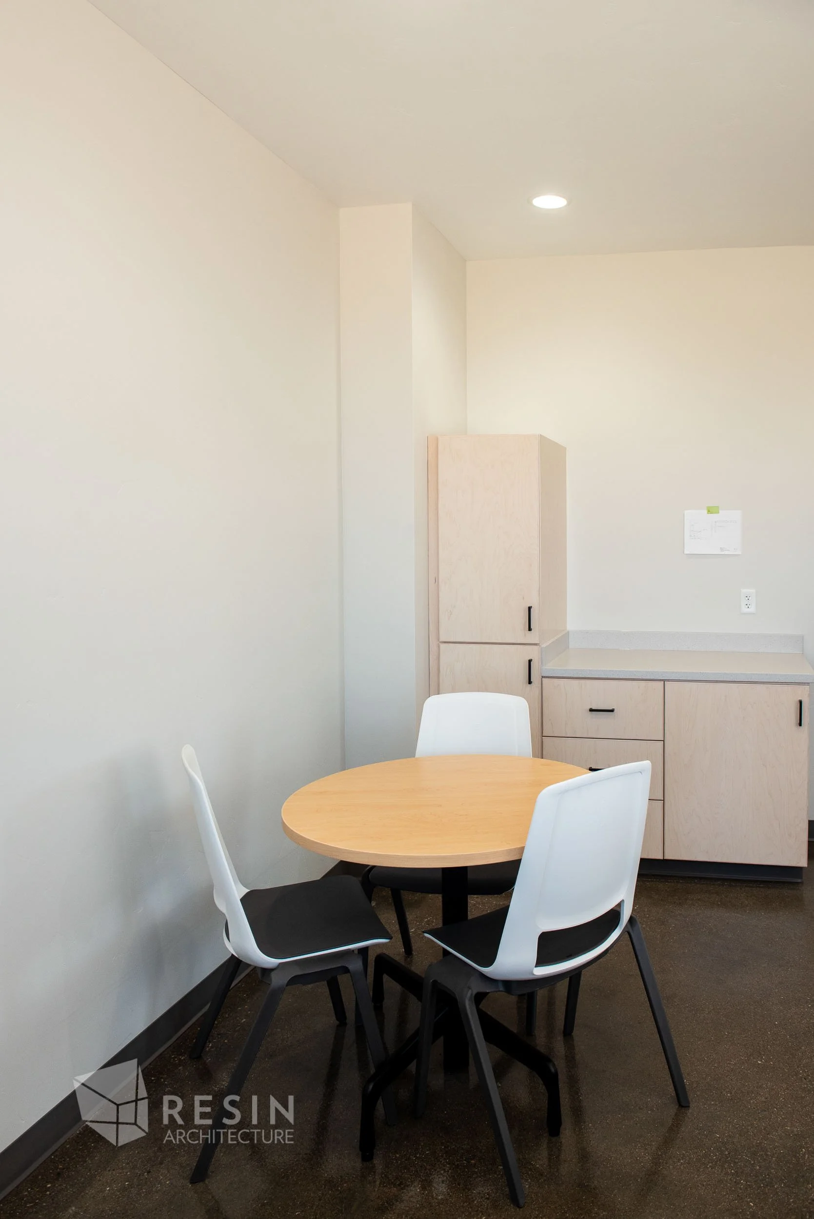Empty round wooden table with four white and black chairs in a room with beige walls and ceiling, gray countertop with cabinets, and a small piece of paper taped to the wall.