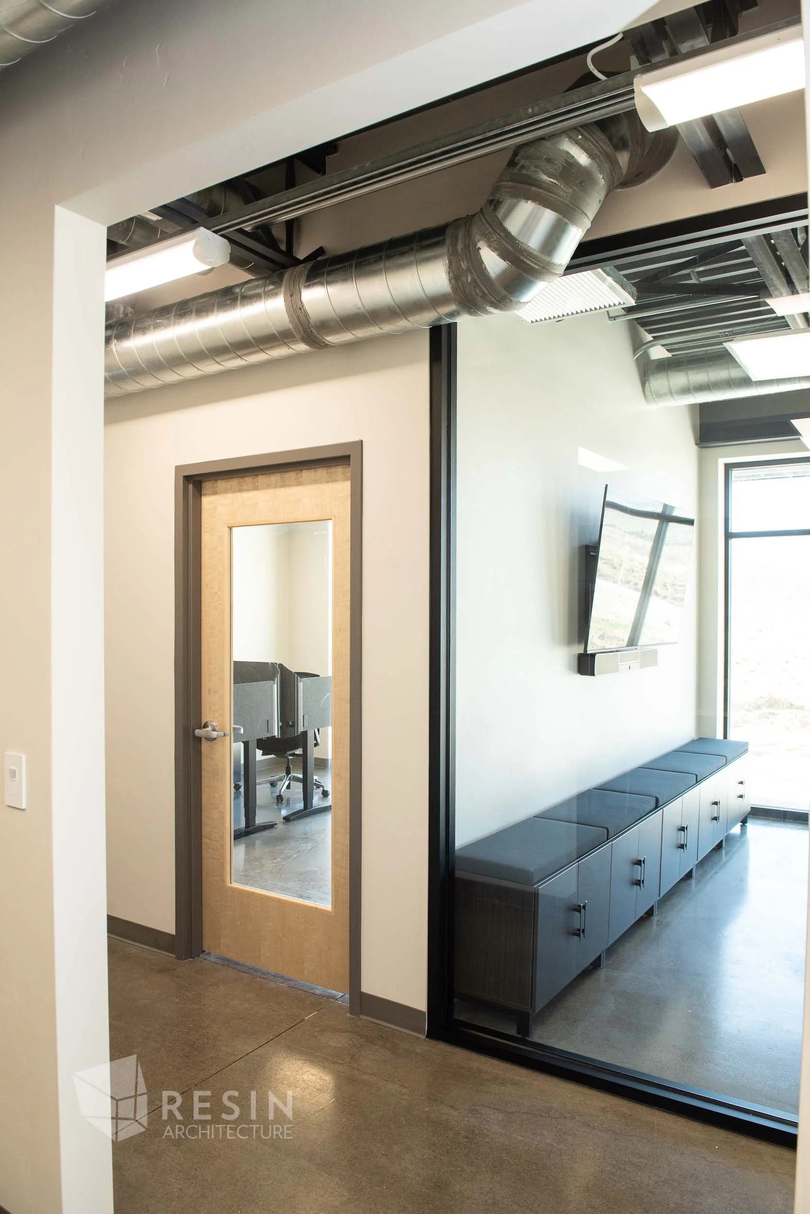 Interior of a modern office space with a door featuring a mirror and a view into a room with desks, a wall-mounted TV, and large windows.