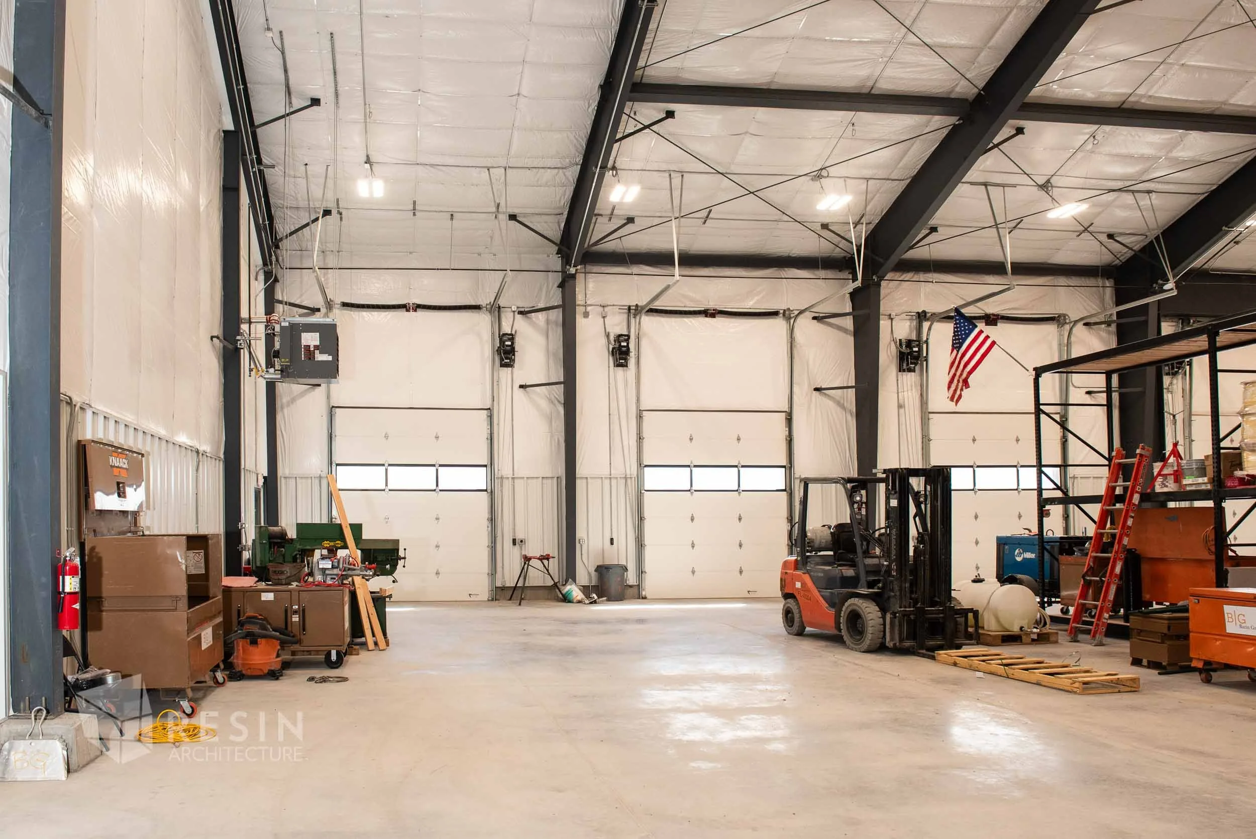 Empty industrial warehouse with large garage doors, storage shelves, a forklift, and equipment.