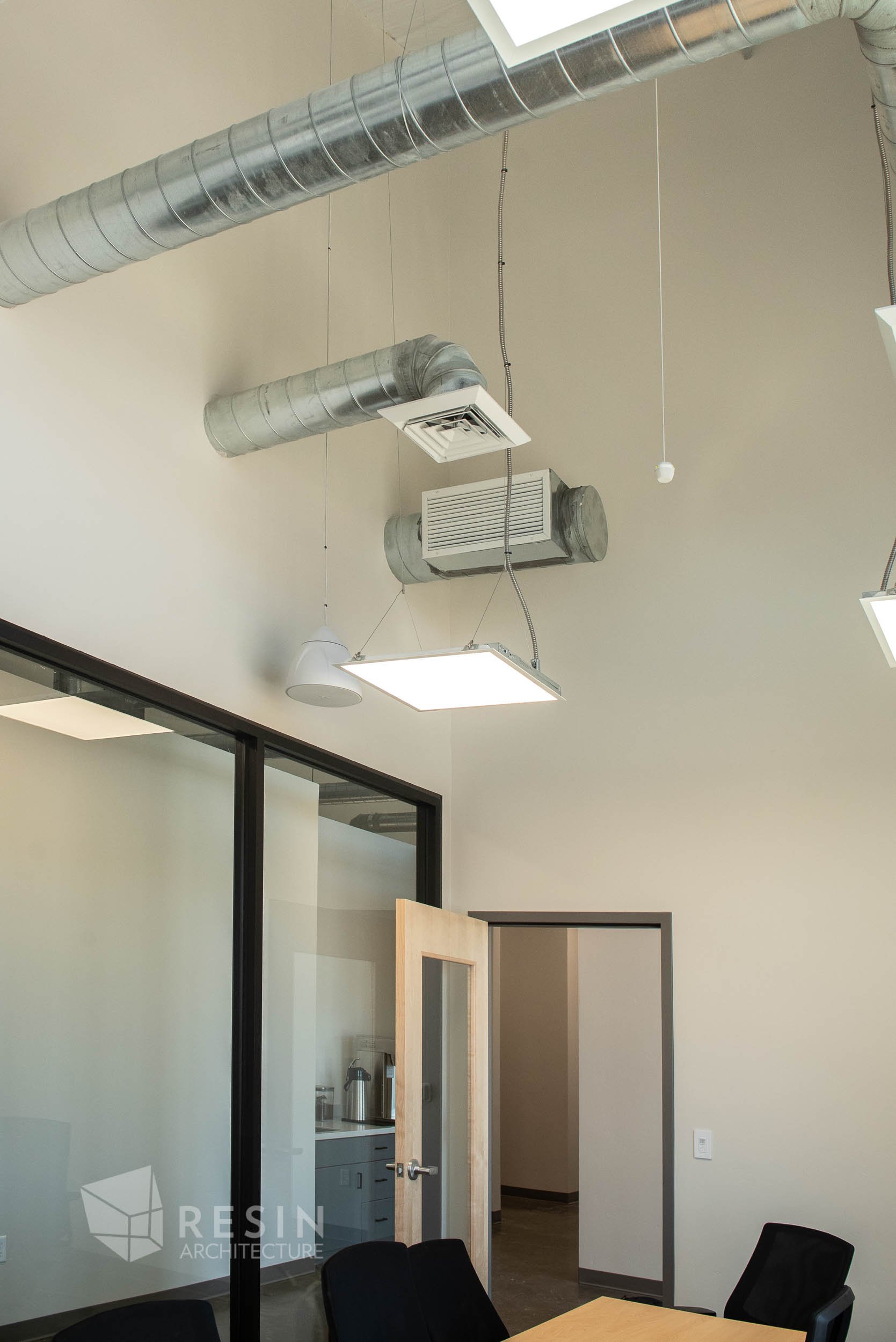 Ceiling with exposed ductwork, air vents, and lighting fixtures in an office conference room with a glass wall, a wooden door, and chairs.