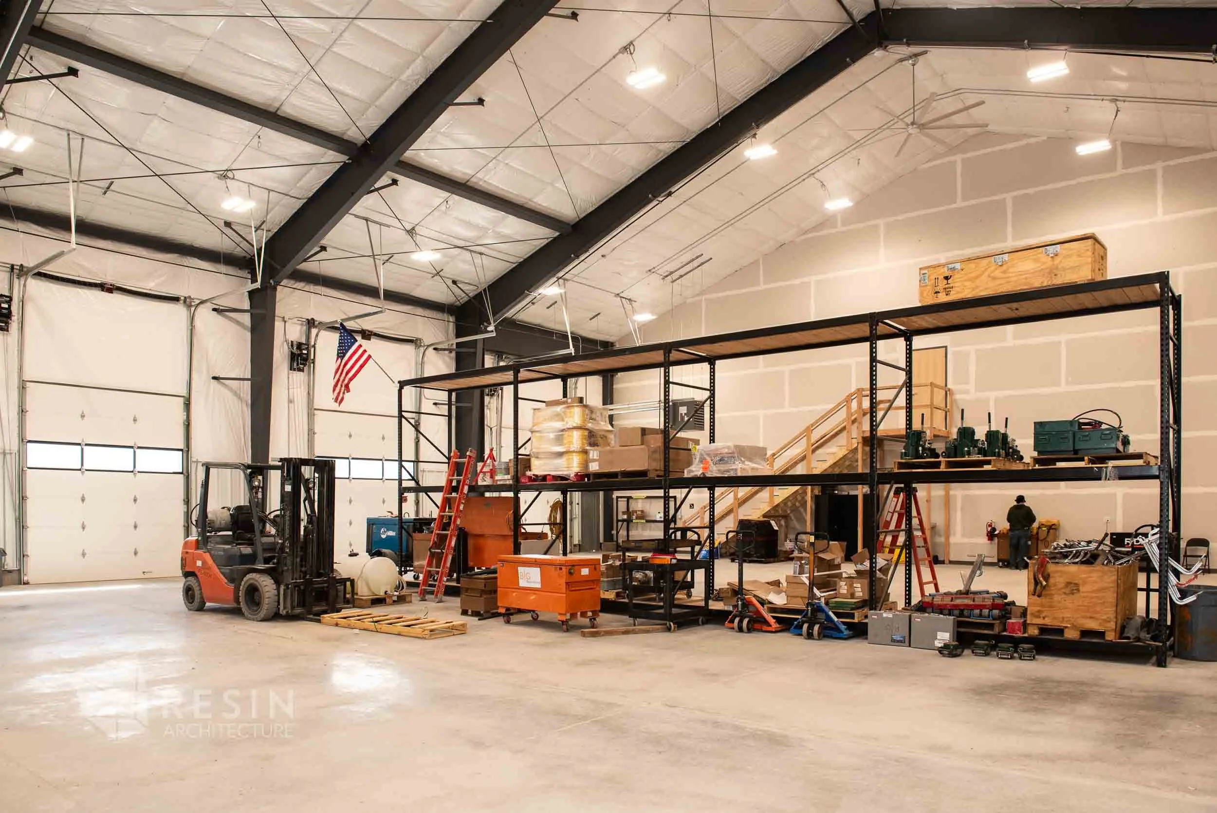 Interior of a warehouse or workshop with a high ceiling, large garage door, metal shelving units filled with tools and equipment, a forklift, and a person working in the background.