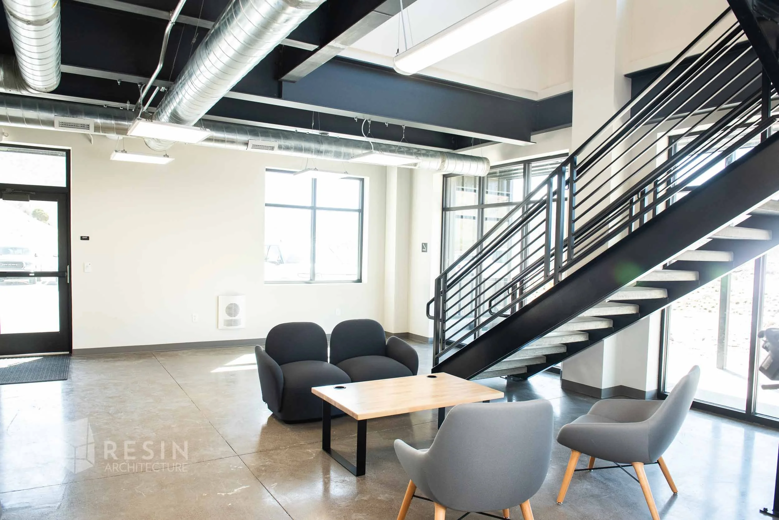 Interior of an office or lobby with modern furniture, large windows, exposed ductwork on the ceiling, a staircase with black railing, and a small seating area with gray and black chairs around a light wood table.