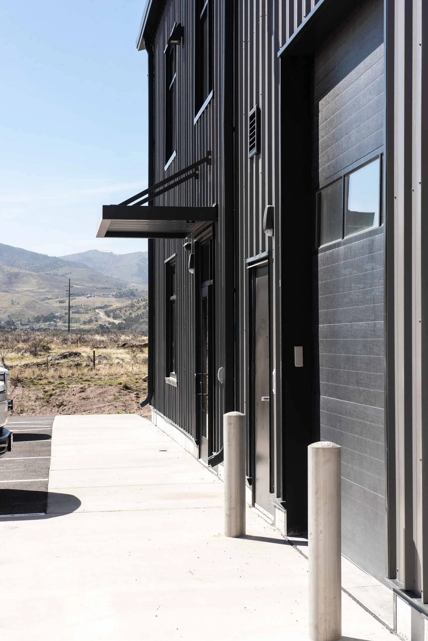 Side view of a modern industrial building with black metallic siding, a parking lot with cars, and mountains in the background.