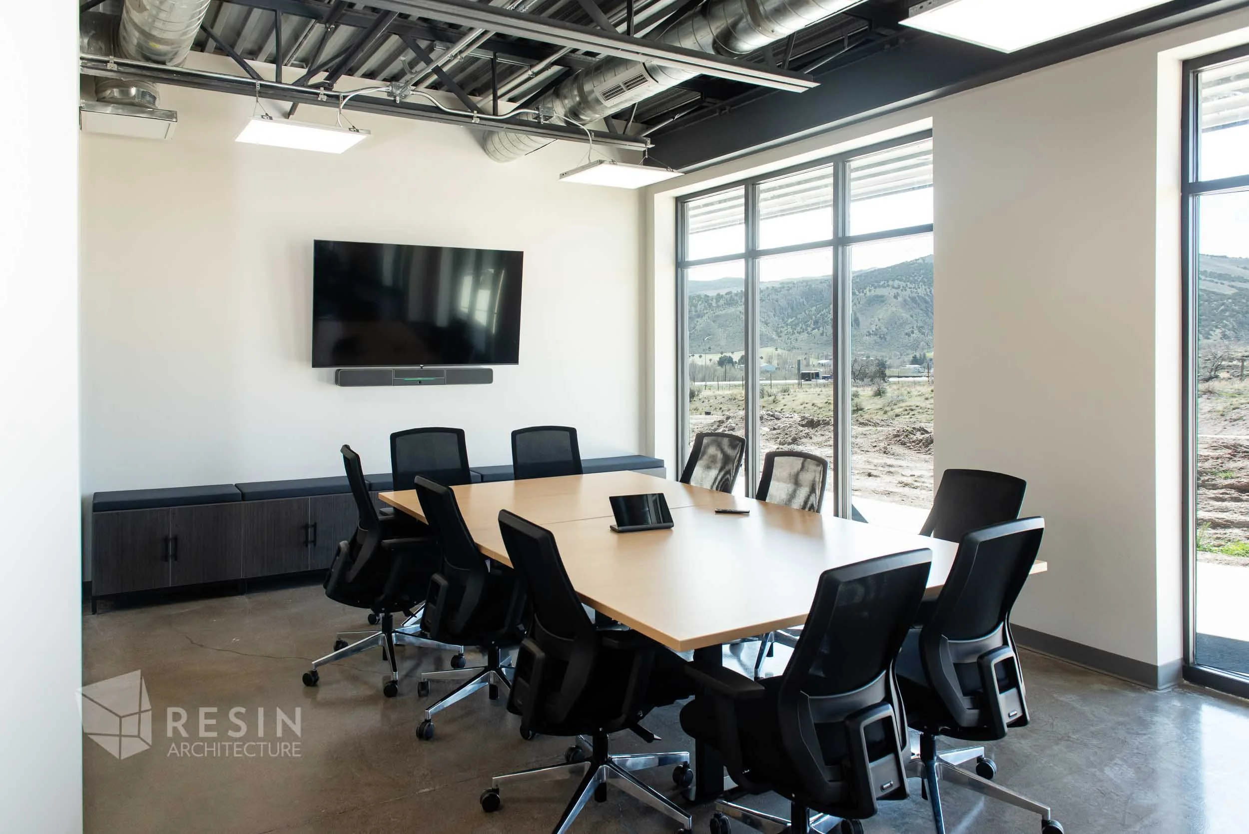 Modern conference room with a large wooden table, black office chairs, a wall-mounted TV, a soundbar below it, large windows showing a mountain landscape, and an exposed ceiling with ductwork.