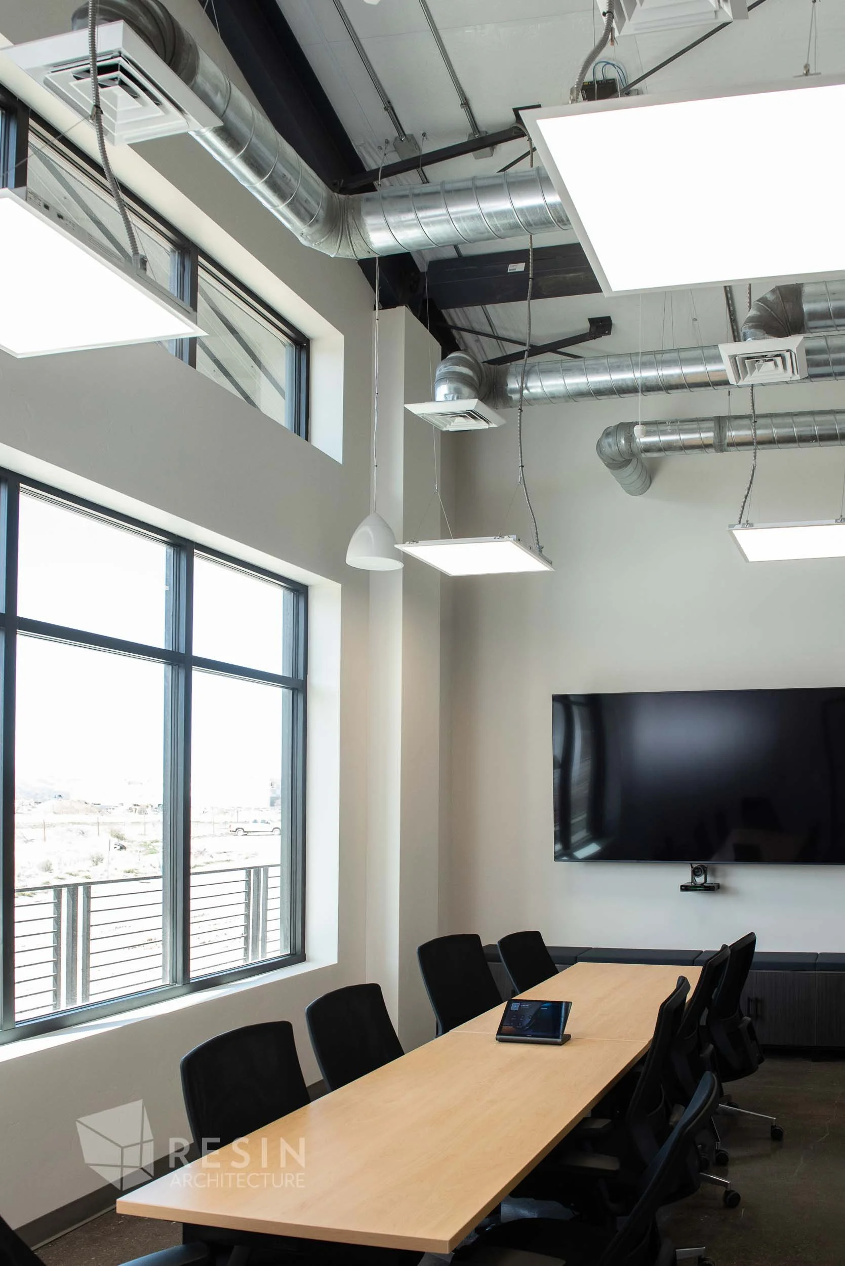 Modern conference room with a long wooden table, black chairs, a large flat-screen TV mounted on the wall, and natural light coming through big windows.