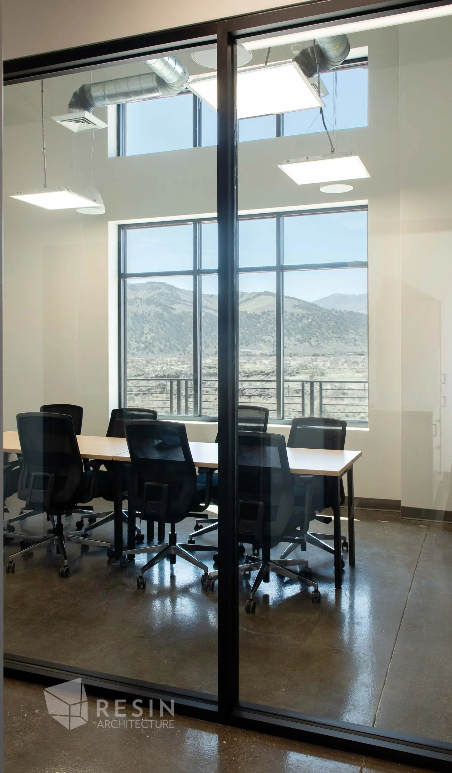 Modern conference room seen through glass wall with black frame, featuring a meeting table, black chairs, large windows with mountain view, and ceiling light fixtures.