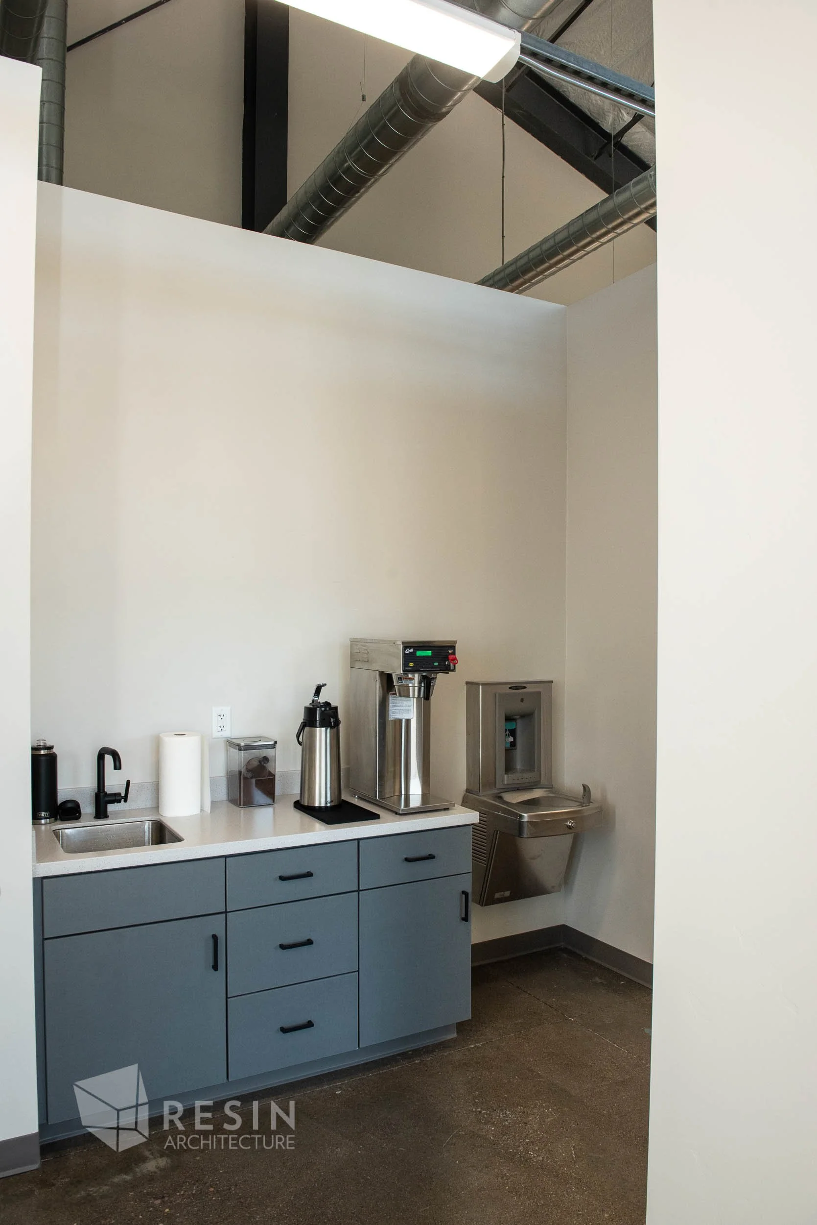 Small kitchenette with a sink, paper towel, hot water dispenser, coffee brewer, and water fountain in an office or break room setting with white walls and industrial ceiling ducts.