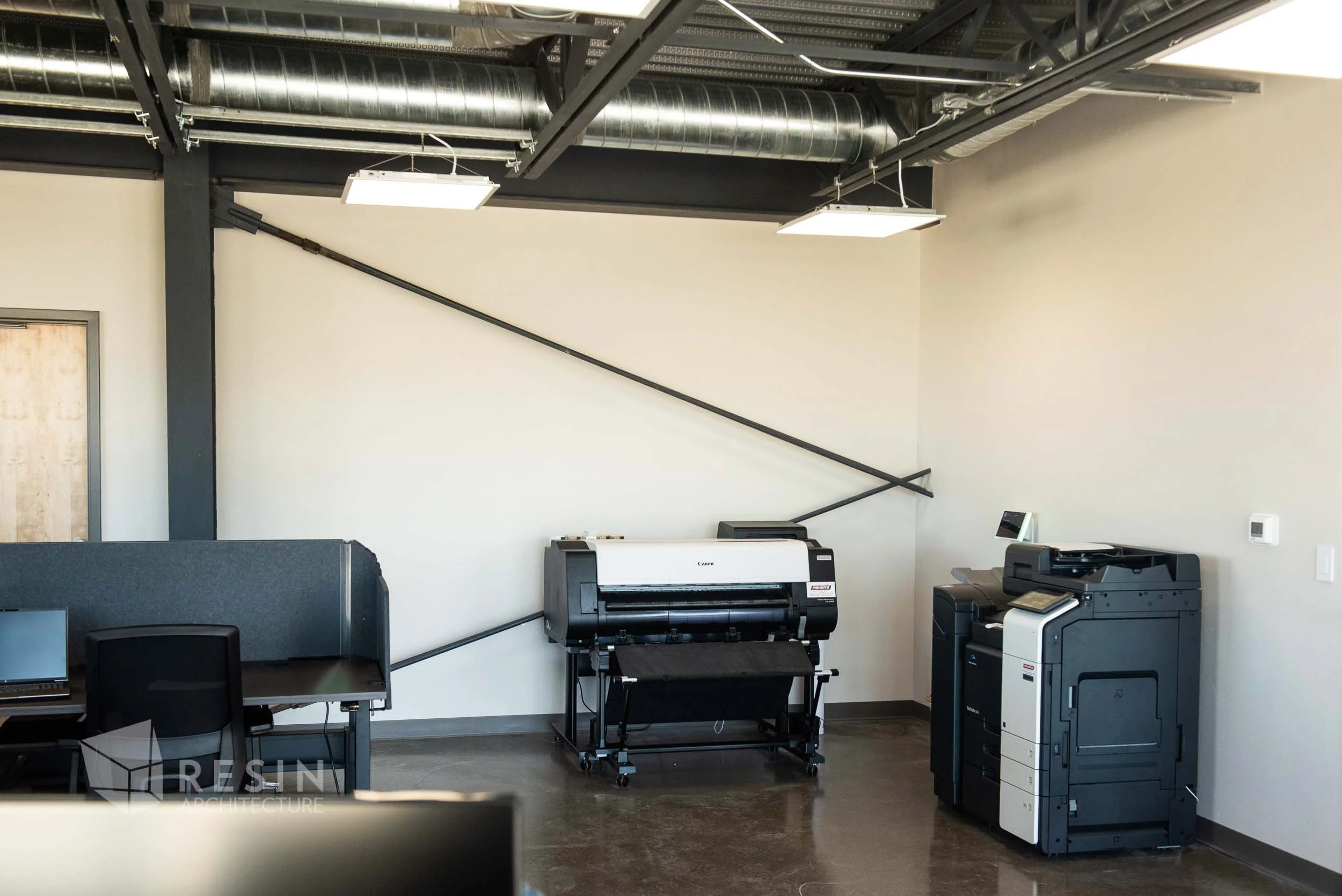 Office space with two black and white printers, a desk with a computer and chair, and exposed ductwork and piping on the ceiling.