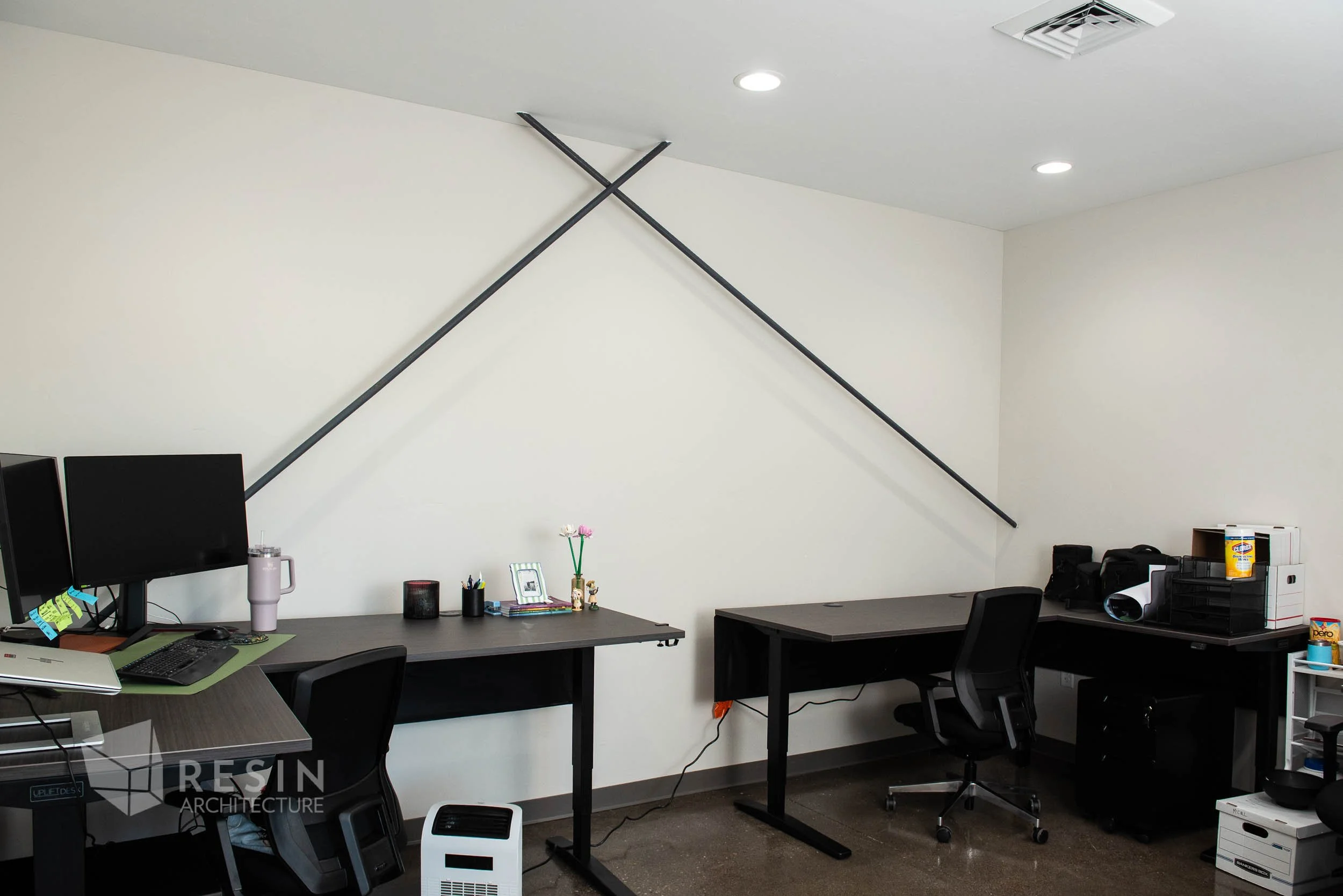 Office with three desks, computers, office chair, and artistic wall decor with black diagonal rods crossing each other.