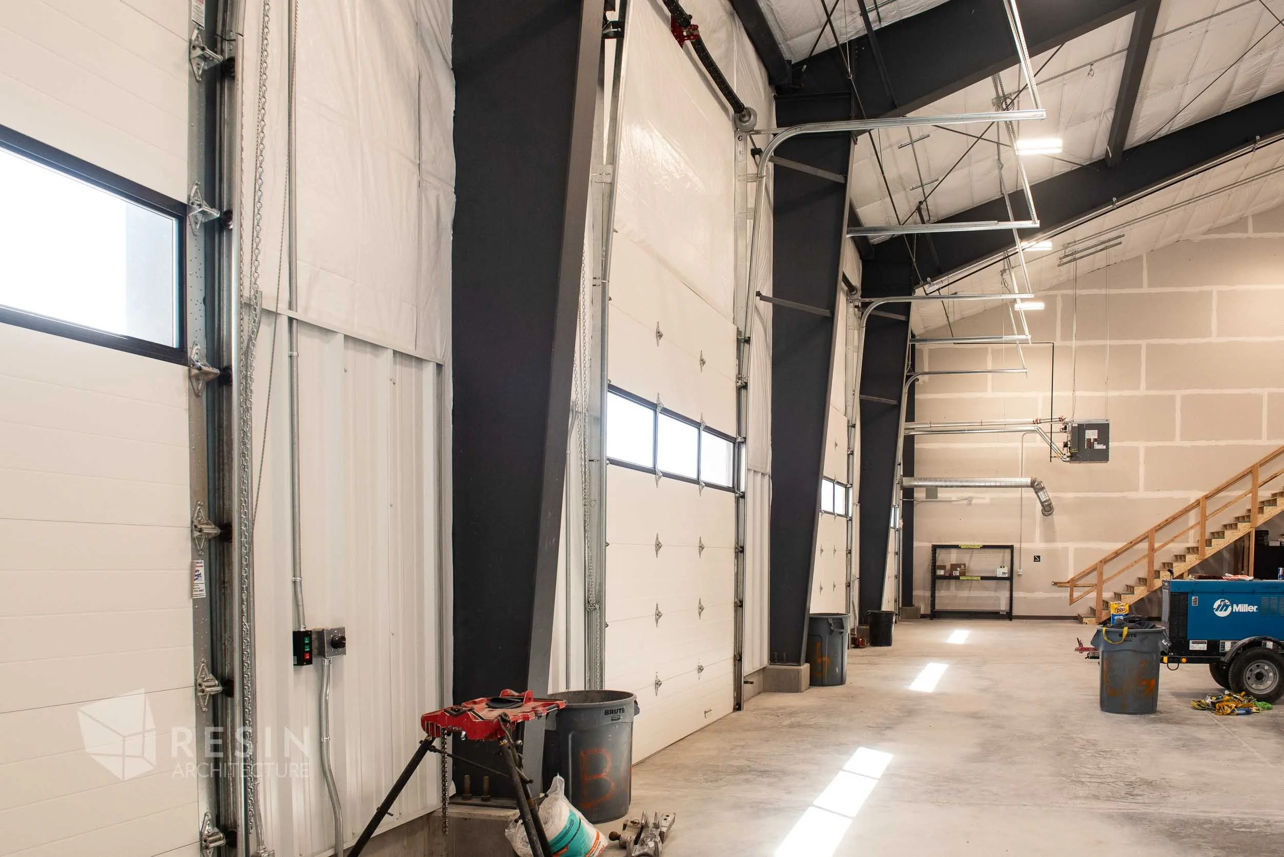 Interior view of a large industrial building with multiple garage doors, a staircase, and construction tools and equipment scattered on the concrete floor.