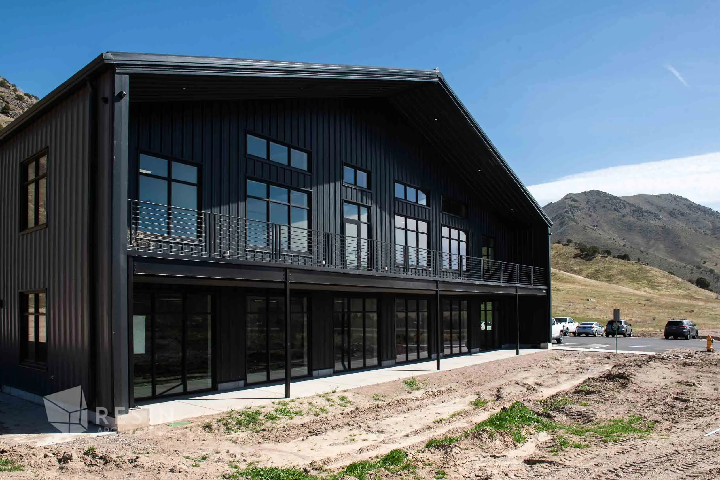 Modern black metal building with large windows and a balcony, situated in a mountainous area with a parking lot and cars nearby.
