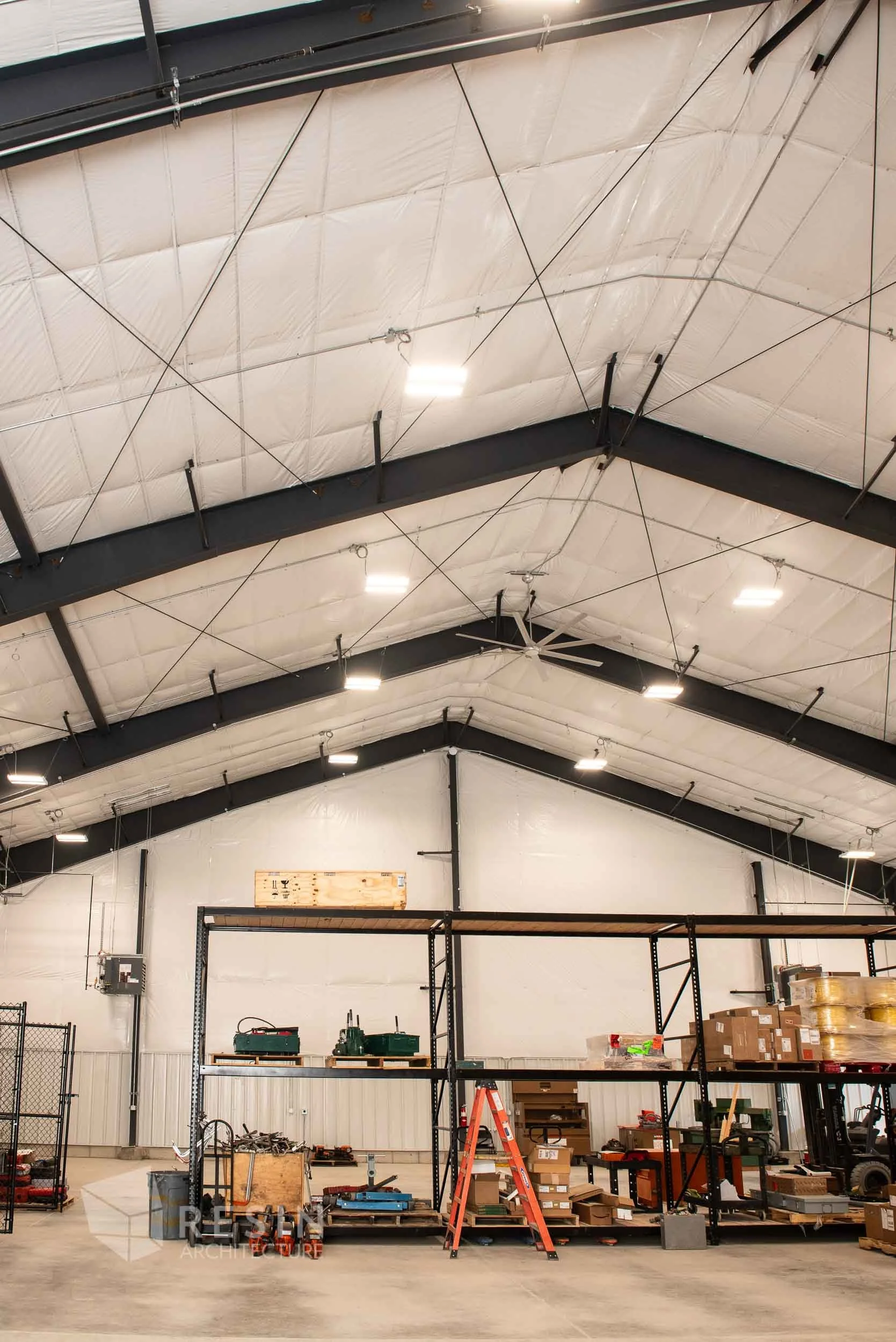 The interior of a warehouse or industrial space with high ceilings, exposed beams, and insulation, featuring metal shelving units with boxes, tools, and equipment, and a red ladder.