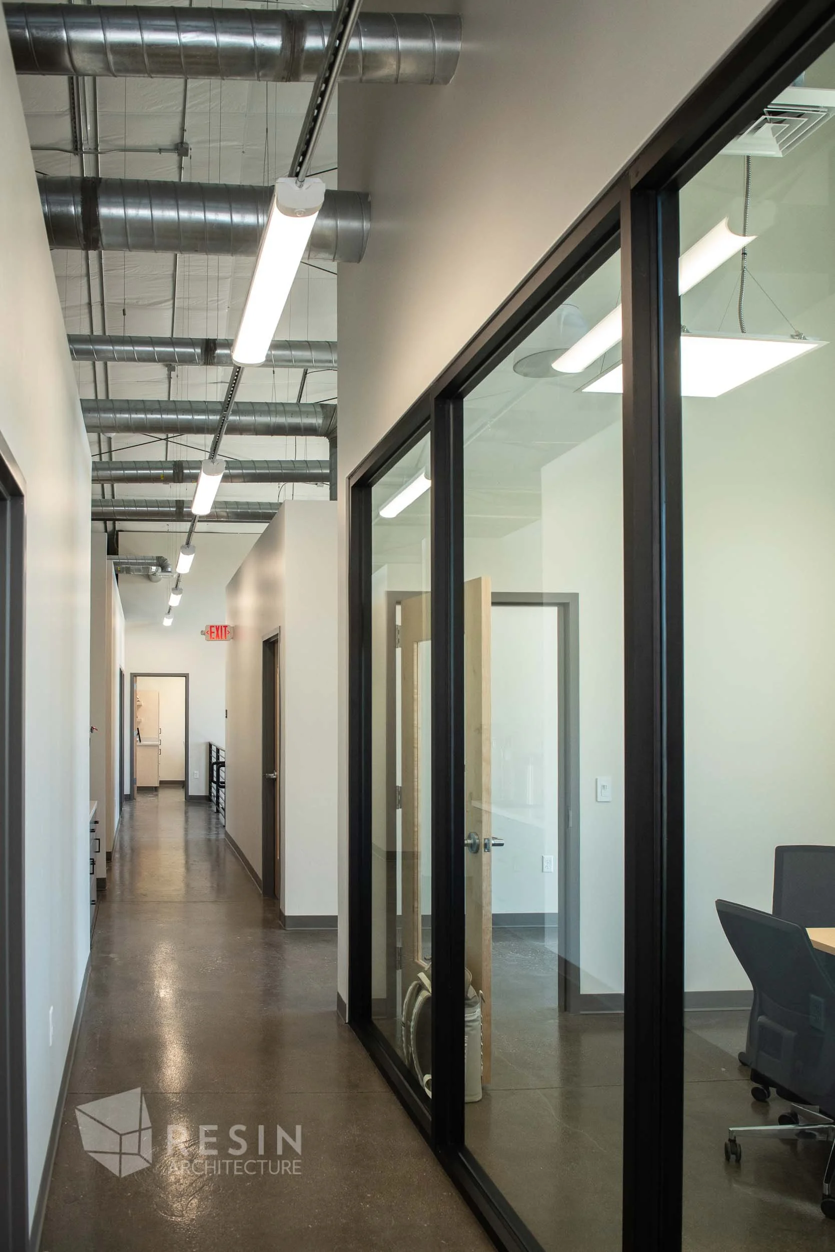 An office hallway with glass-walled offices on the right, exposed ductwork on the ceiling, and fluorescent lights. The floor is polished concrete and there is an exit sign at the far end.