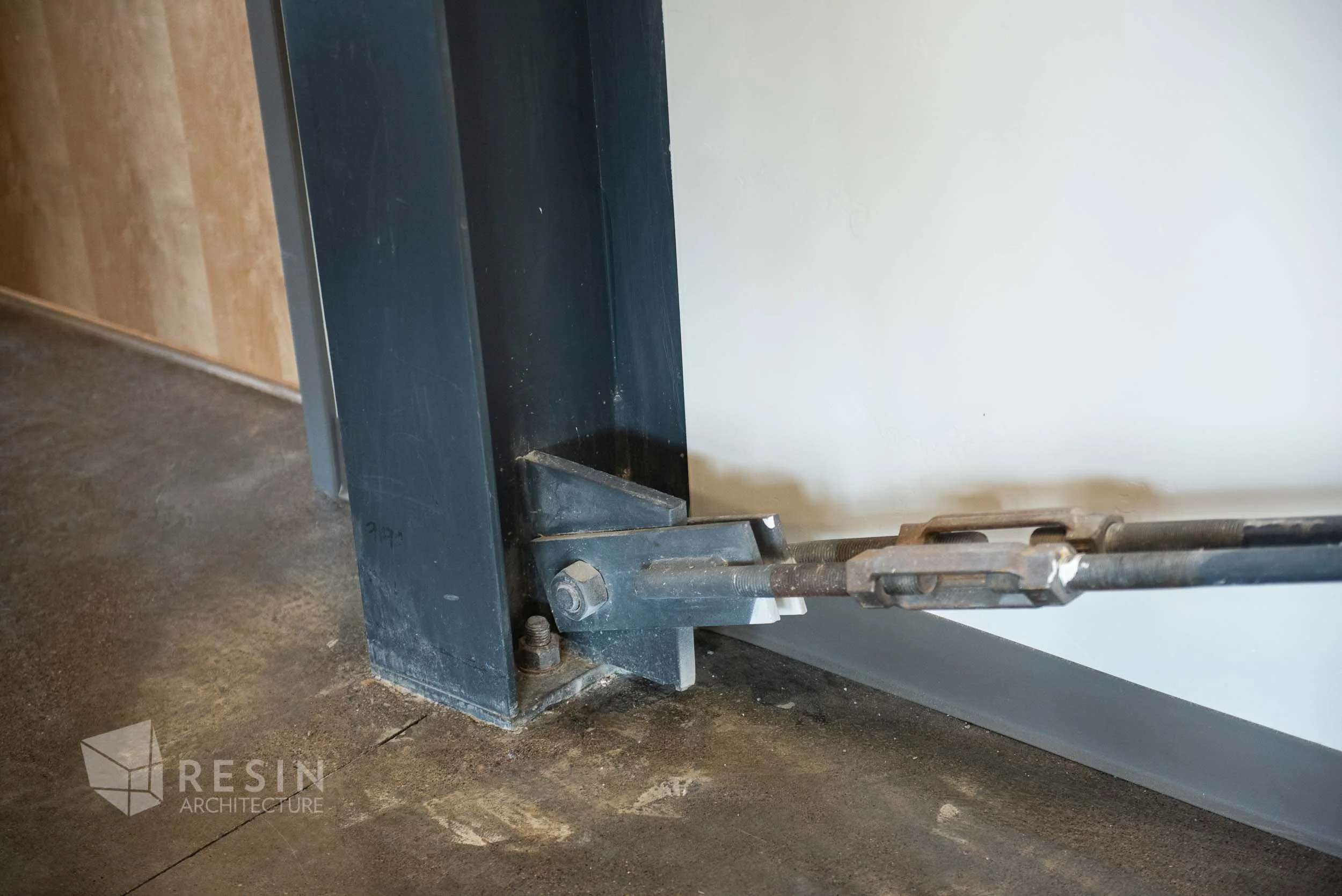 Close-up of a metal support beam and anchoring hardware at the base of a wall, with a concrete floor.