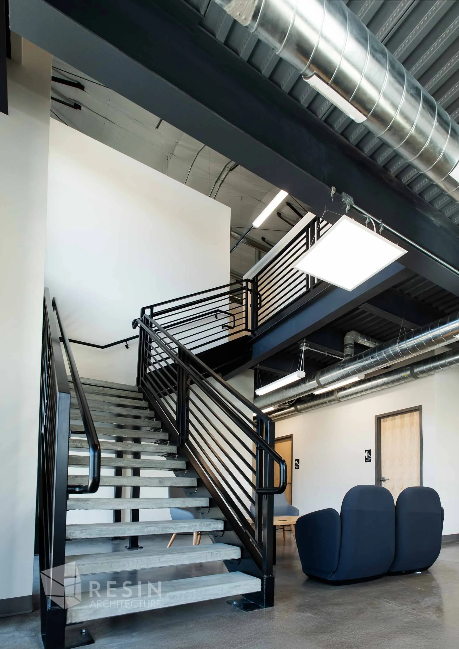 Modern industrial interior with metal staircase, waiting area with black sofa, doors labeled for restrooms, exposed ductwork, and ceiling lighting.