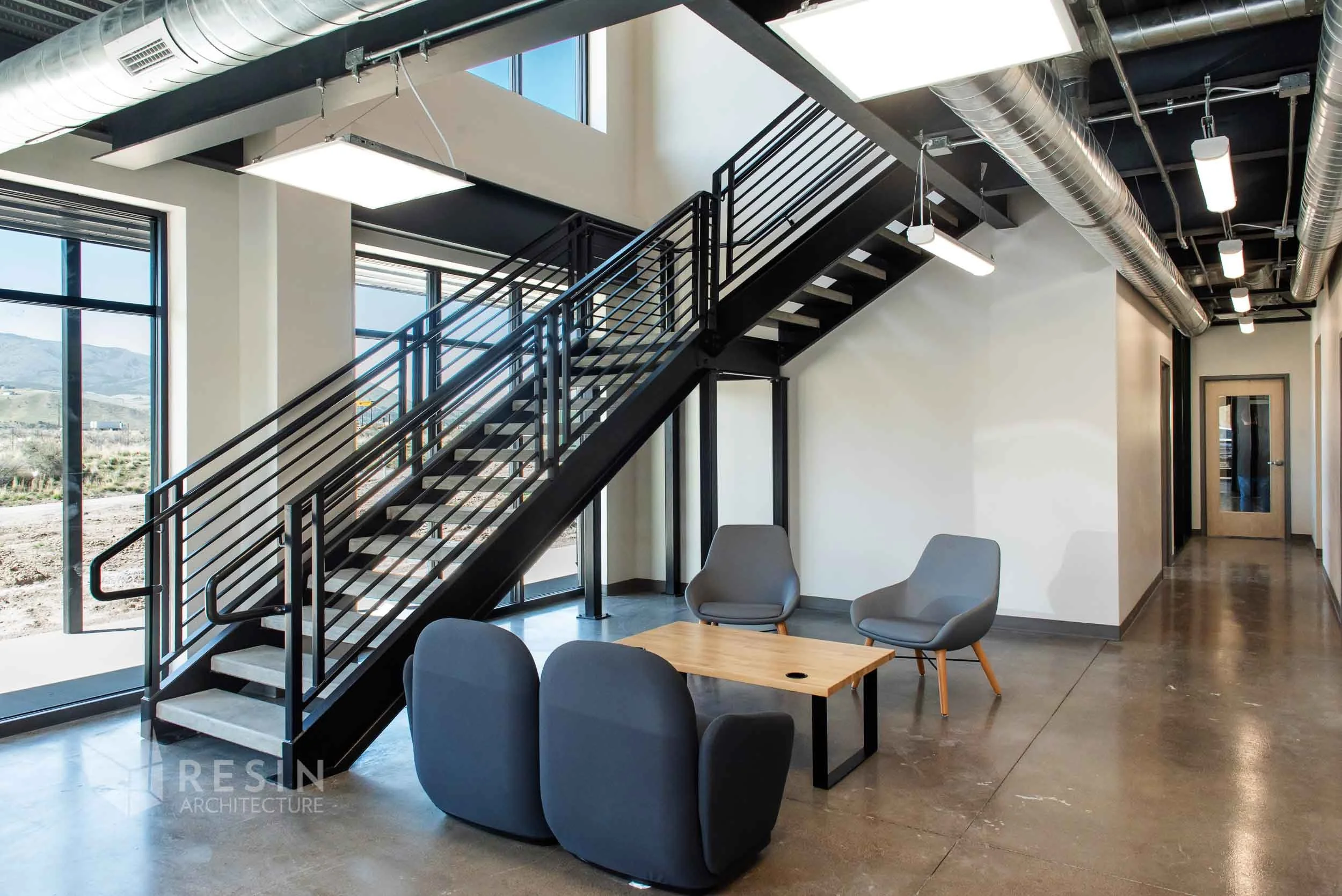 Interior of an office space with a black metal staircase, large windows showing a mountainous landscape, and modern gray chairs around a small wooden table.