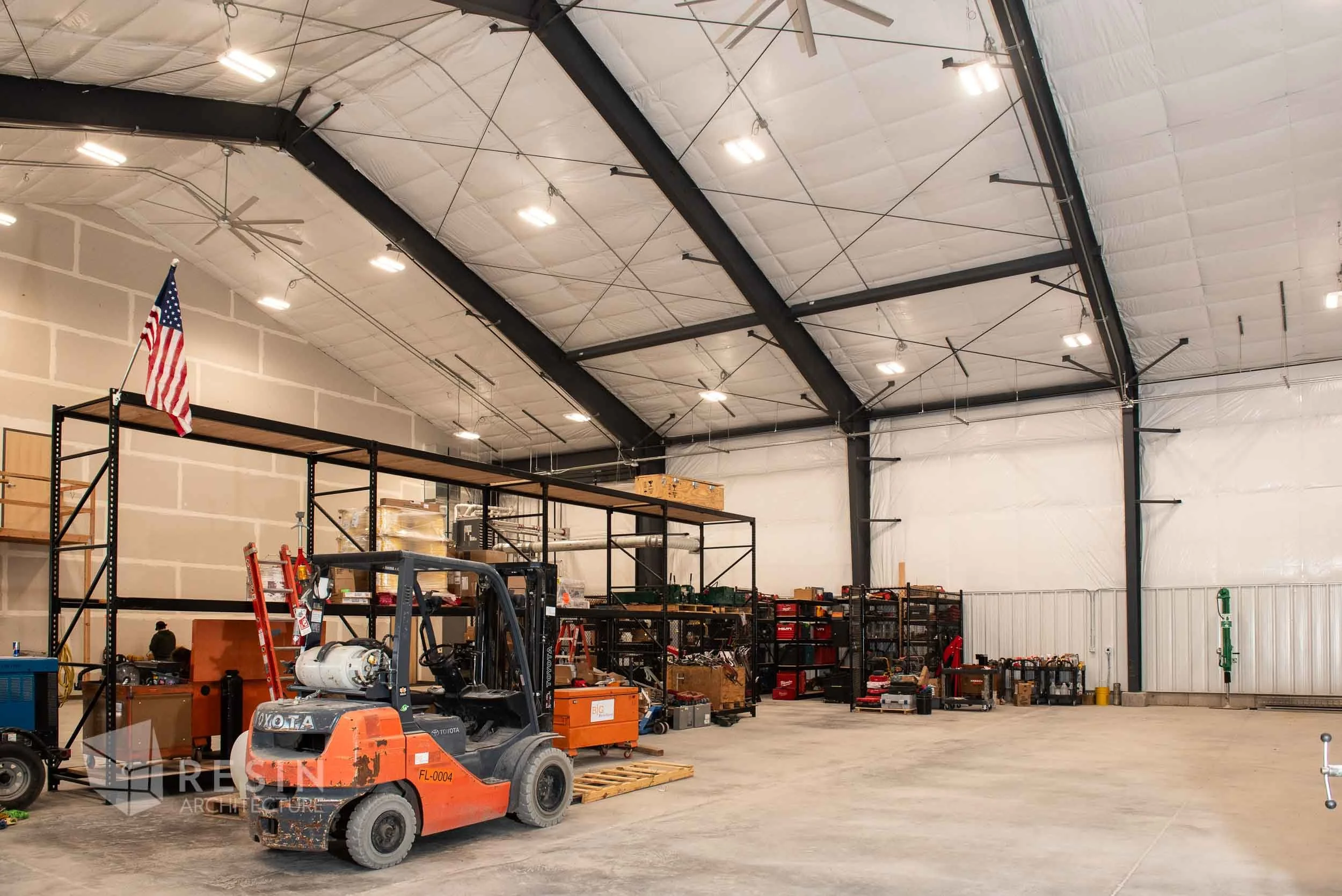 Interior view of a warehouse with high ceilings, industrial lighting, metal shelving units filled with tools and equipment, and a forklift parked on the concrete floor.
