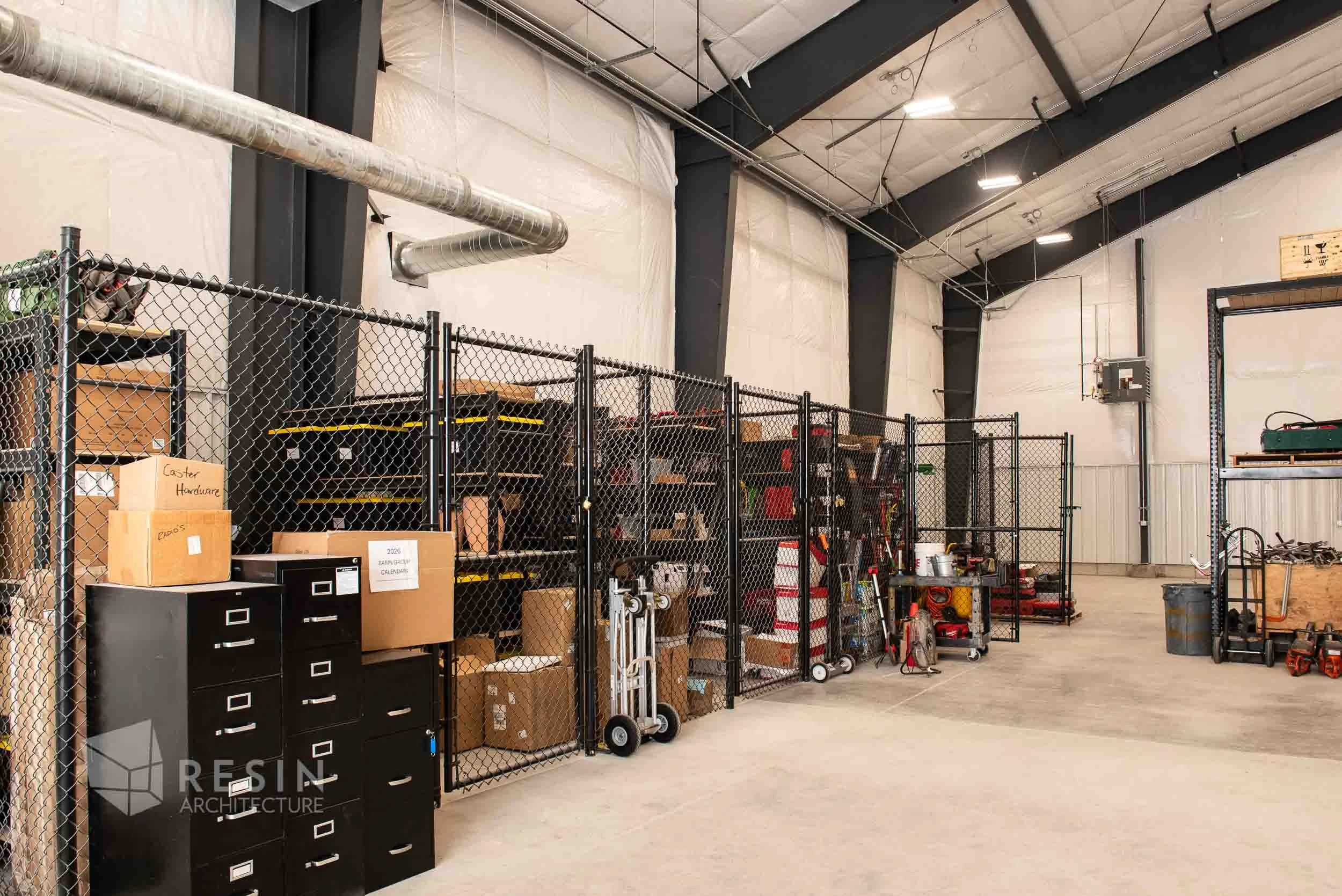 Inside a warehouse with metal shelves and storage boxes, orange safety cones, and various tools and equipment, enclosed by a black wire fence.