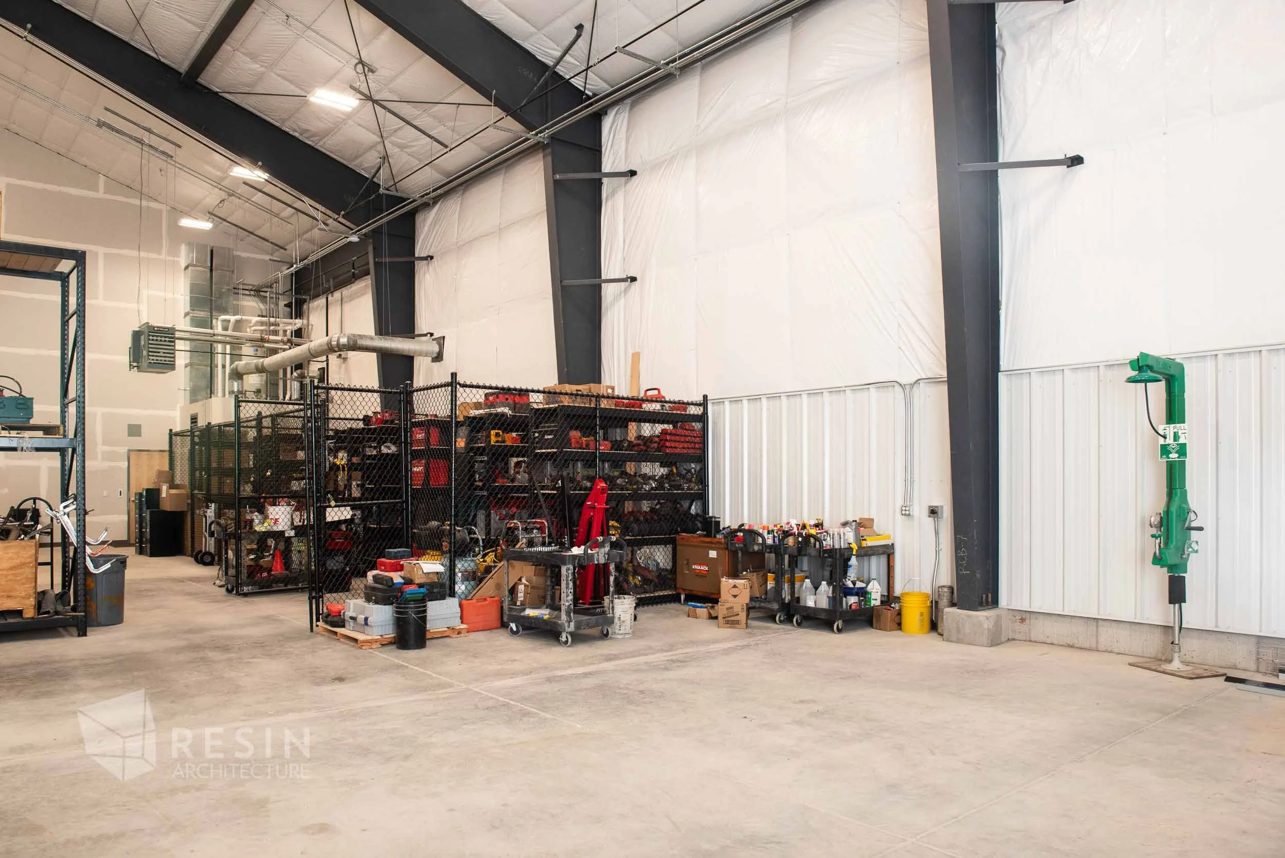Industrial warehouse storage area with tools, equipment, and supplies organized on black metal shelves and carts, concrete floor, and high ceiling.