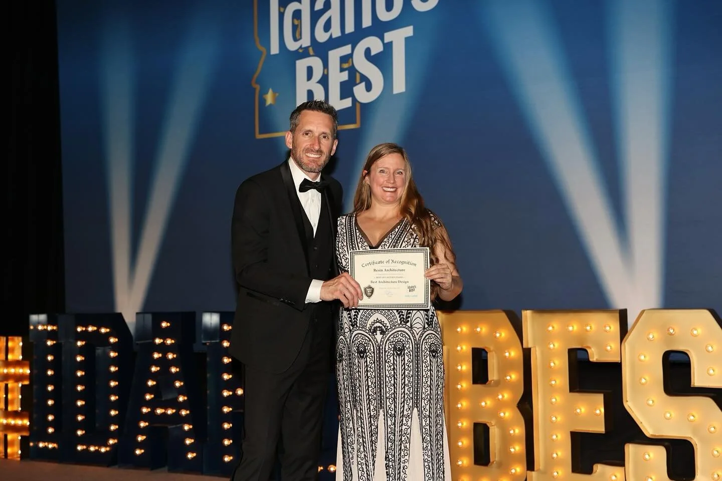 A man and Jamee Moulton from Resin Architecture dressed in formal attire, standing on stage, holding a certificate of recognition, smiling, in front of illuminated large letters and a backdrop with the words 'Idaho's Best.'