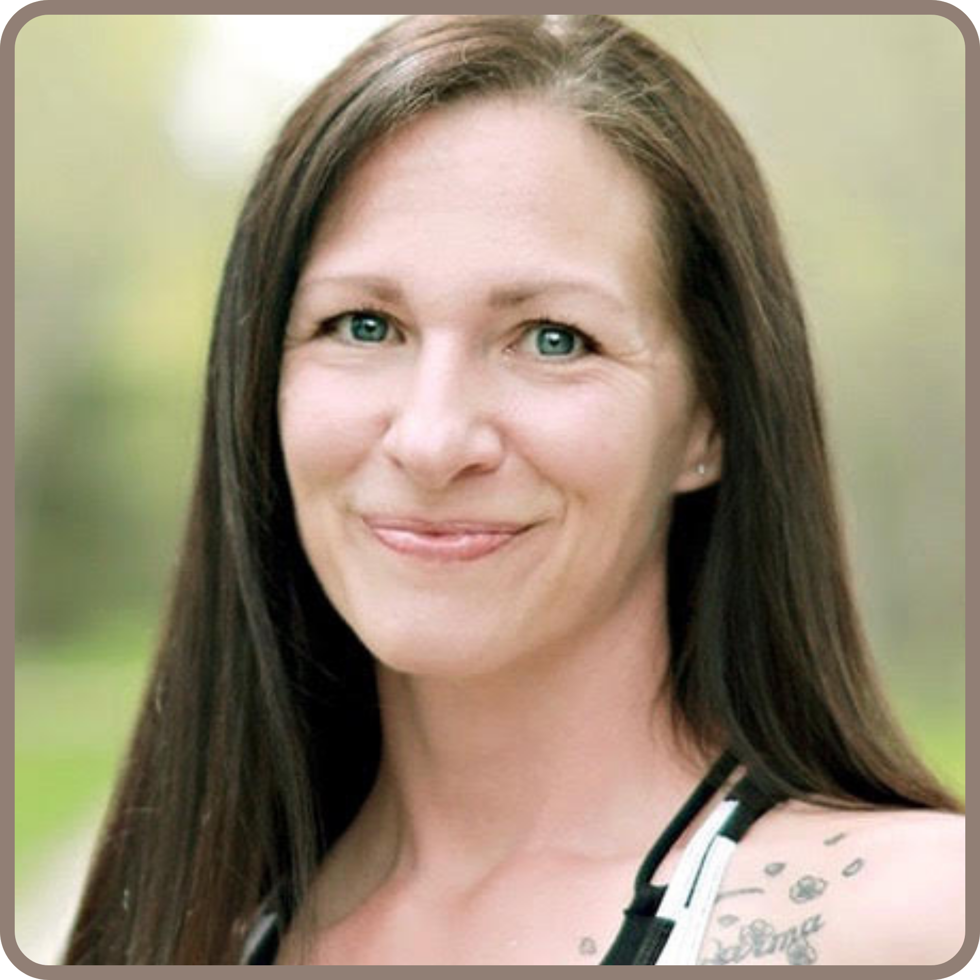 Close-up of a woman with long brown hair and blue eyes, smiling, wearing a polka-dot shirt with a green and white striped neckline, against a blurred green background.