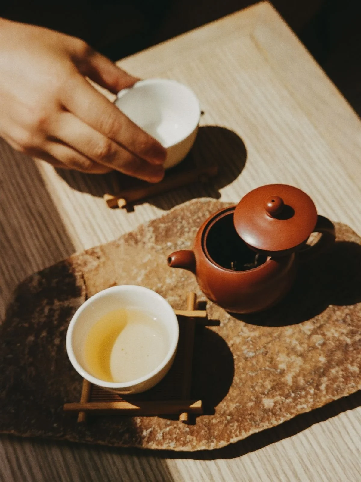 A tea set including a small brown teapot, a white teacup with green tea, and an egg in a cup, placed on a rustic stone slab with a wooden tray underneath. A hand is holding the egg cup, and the setup is on a wooden table.