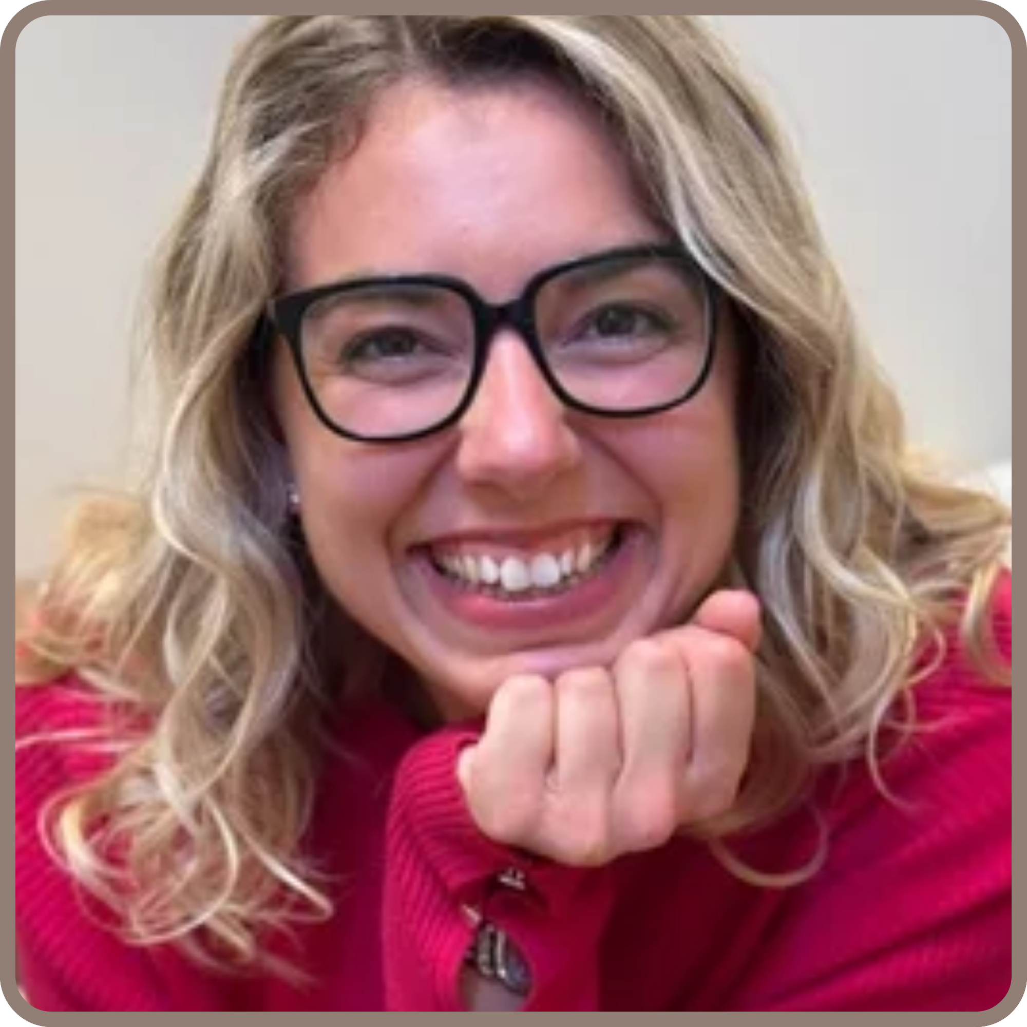 Close-up of a woman with long, wavy blonde hair and glasses, smiling and resting her chin on her hands, wearing a pink sweater.