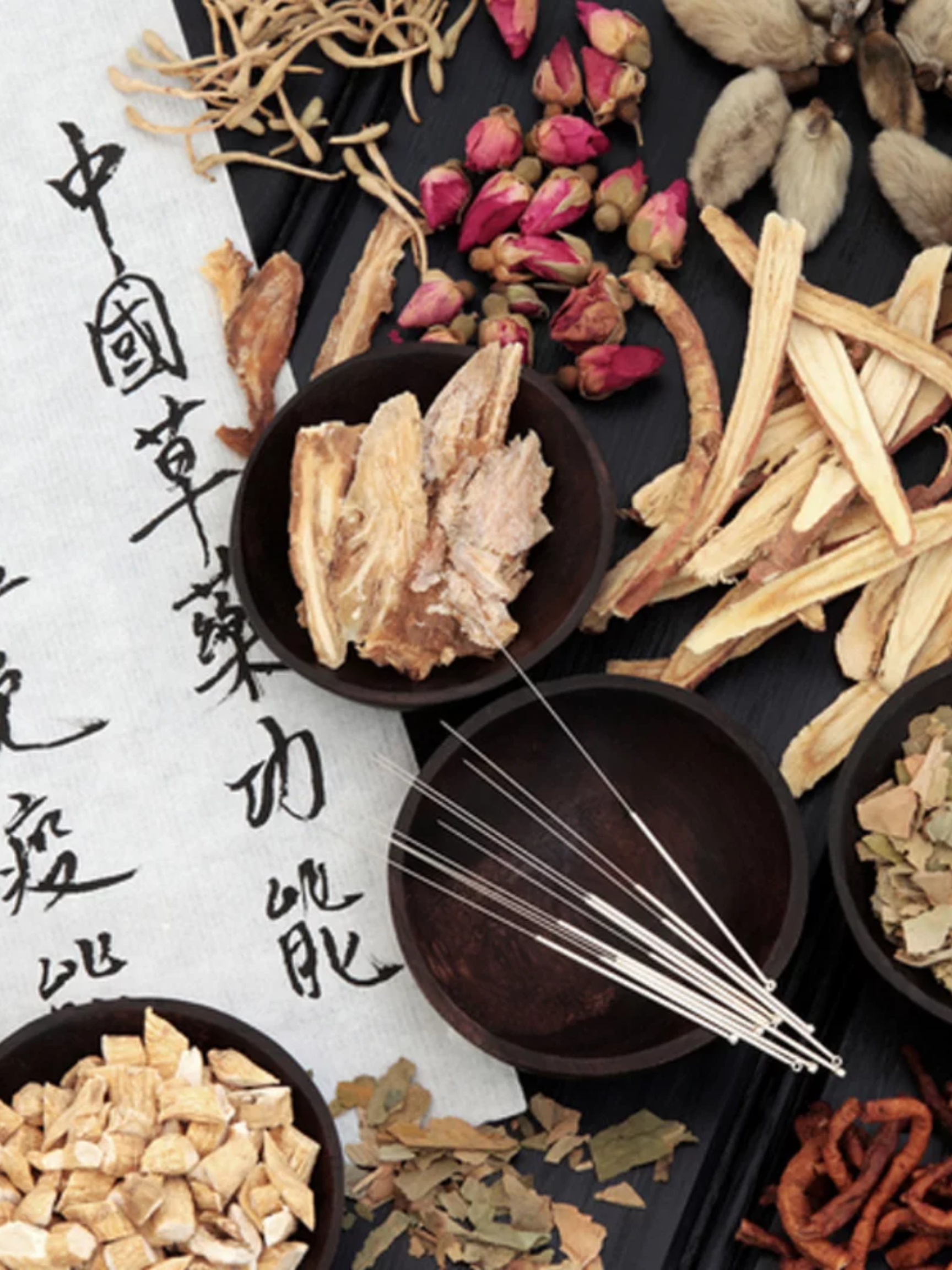Close-up of traditional Asian medicinal ingredients, including dried herbs, roots, and flowers, arranged on a black tray with handwritten Chinese or Japanese characters on a white paper in the background.