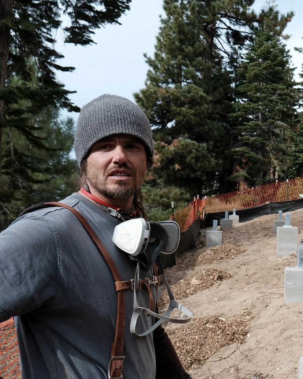 A man in a gray beanie and dark shirt with a respirator mask around his neck is standing outdoors on a dirt hill with construction barriers and trees in the background.