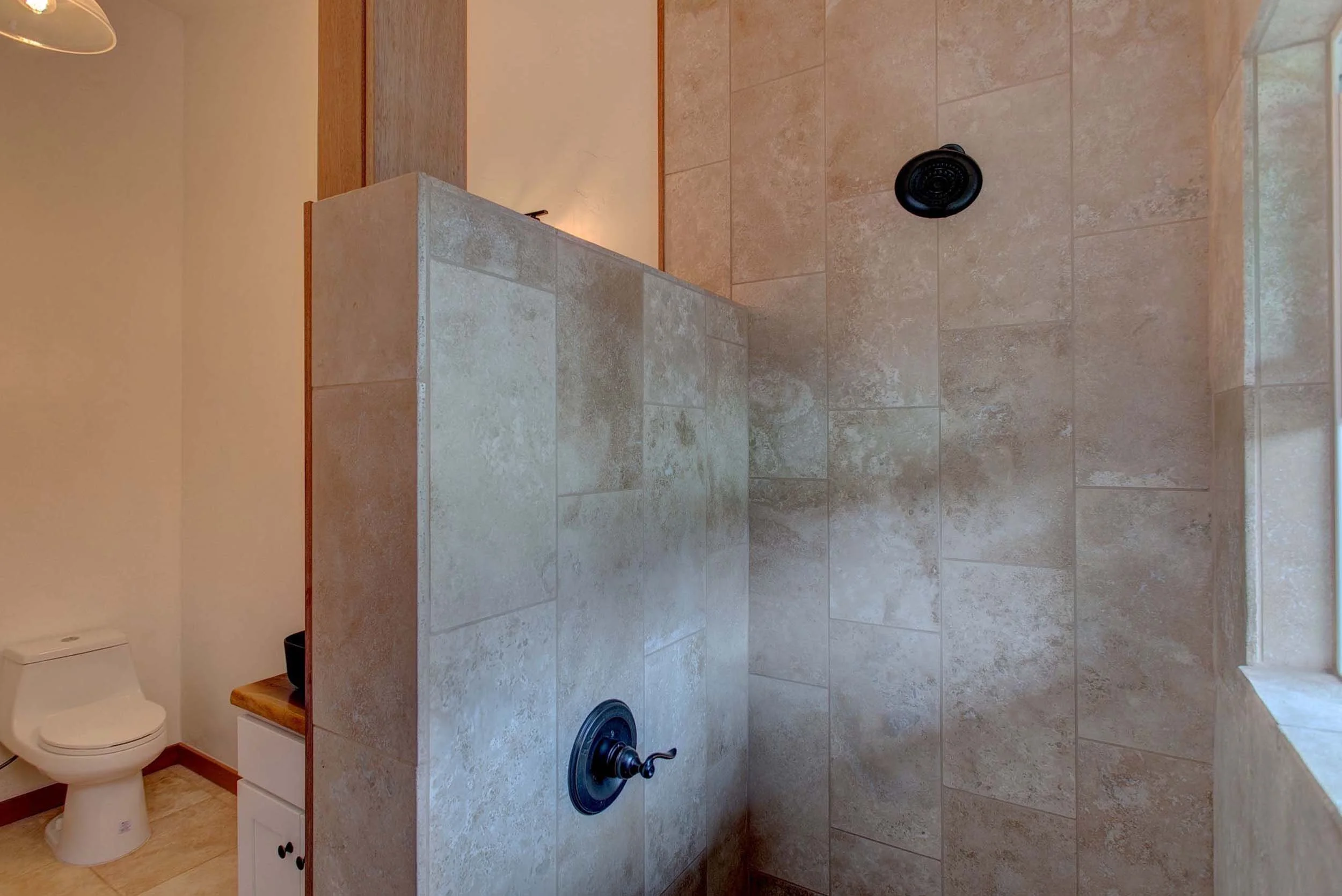A bathroom with a walk-in shower featuring beige tiles, a black showerhead, and a partial view of a toilet and a small cabinet.