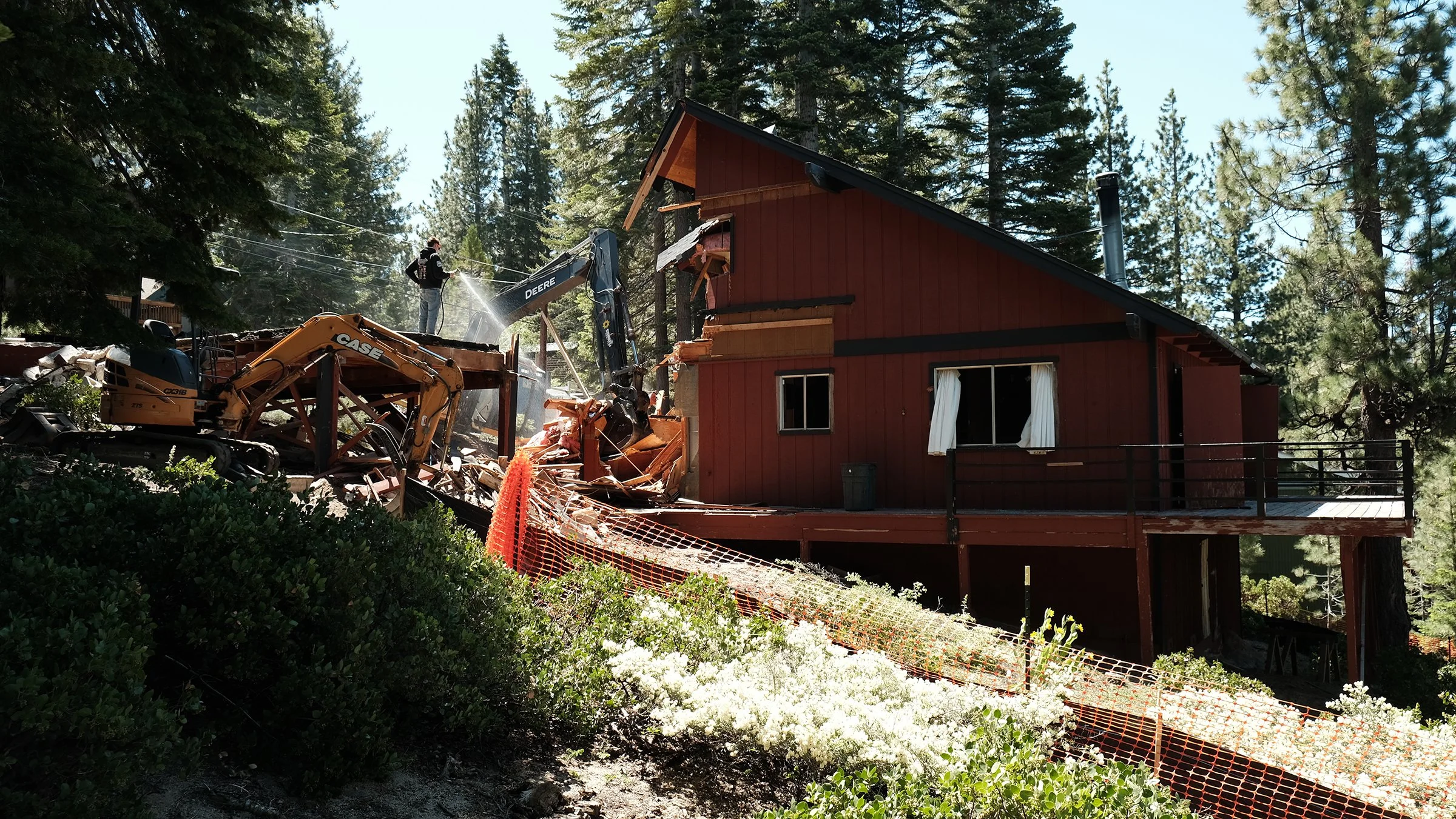 A house being demolished with construction workers and equipment, surrounded by trees and greenery.