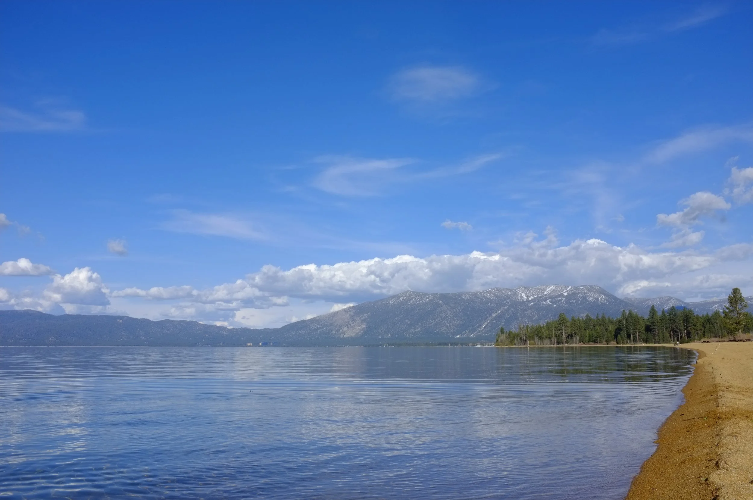 A tranquil lake with calm water reflecting the blue sky and scattered clouds, surrounded by mountains and a sandy shoreline with trees.