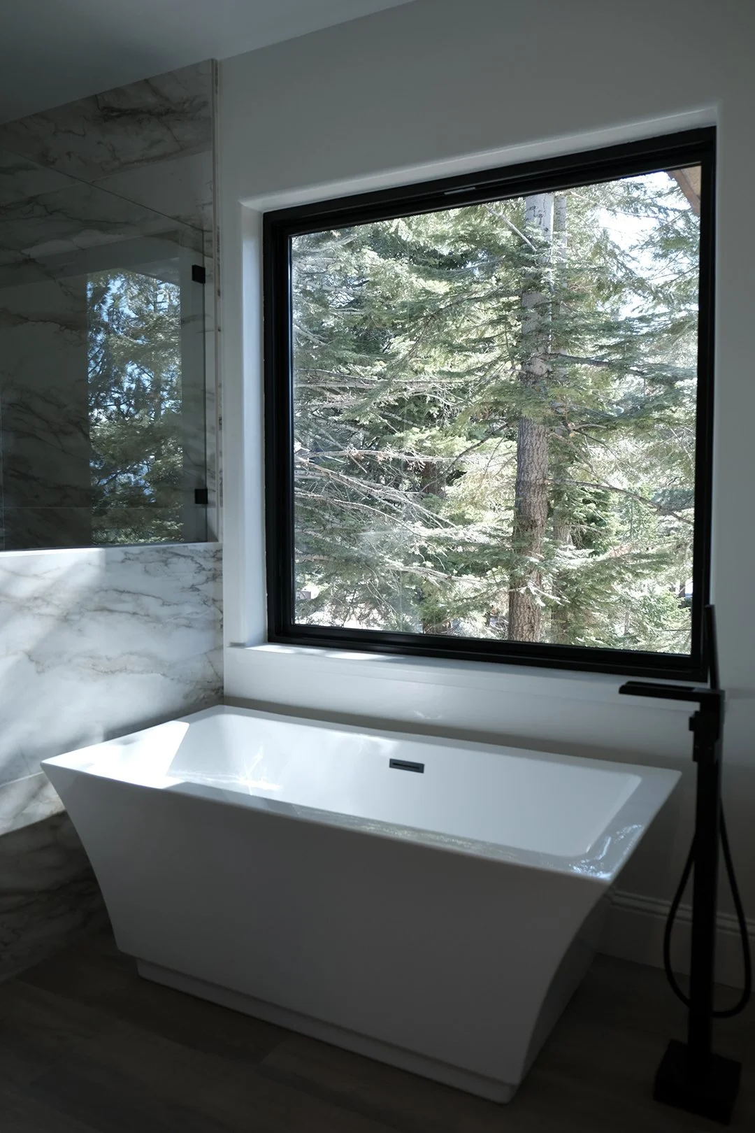 Modern bathroom with a white freestanding bathtub in front of a large window showing trees outside.