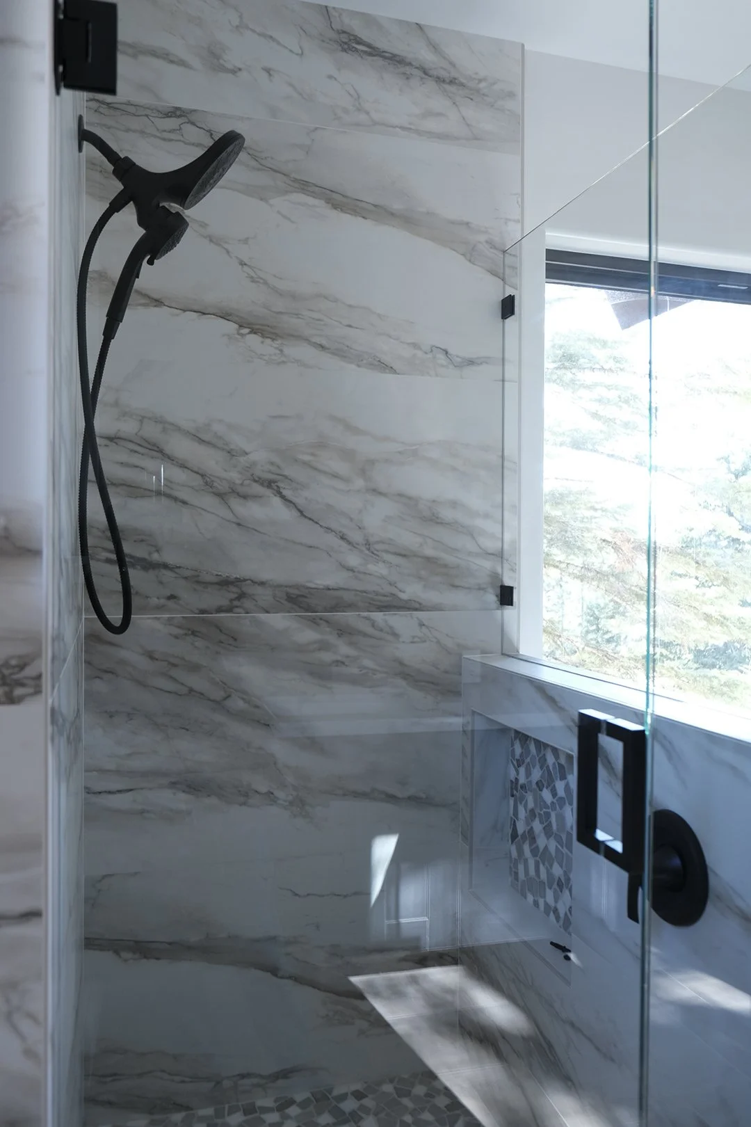 Modern shower with marble walls, a black handheld showerhead, and a glass door, with a window showing trees outside.