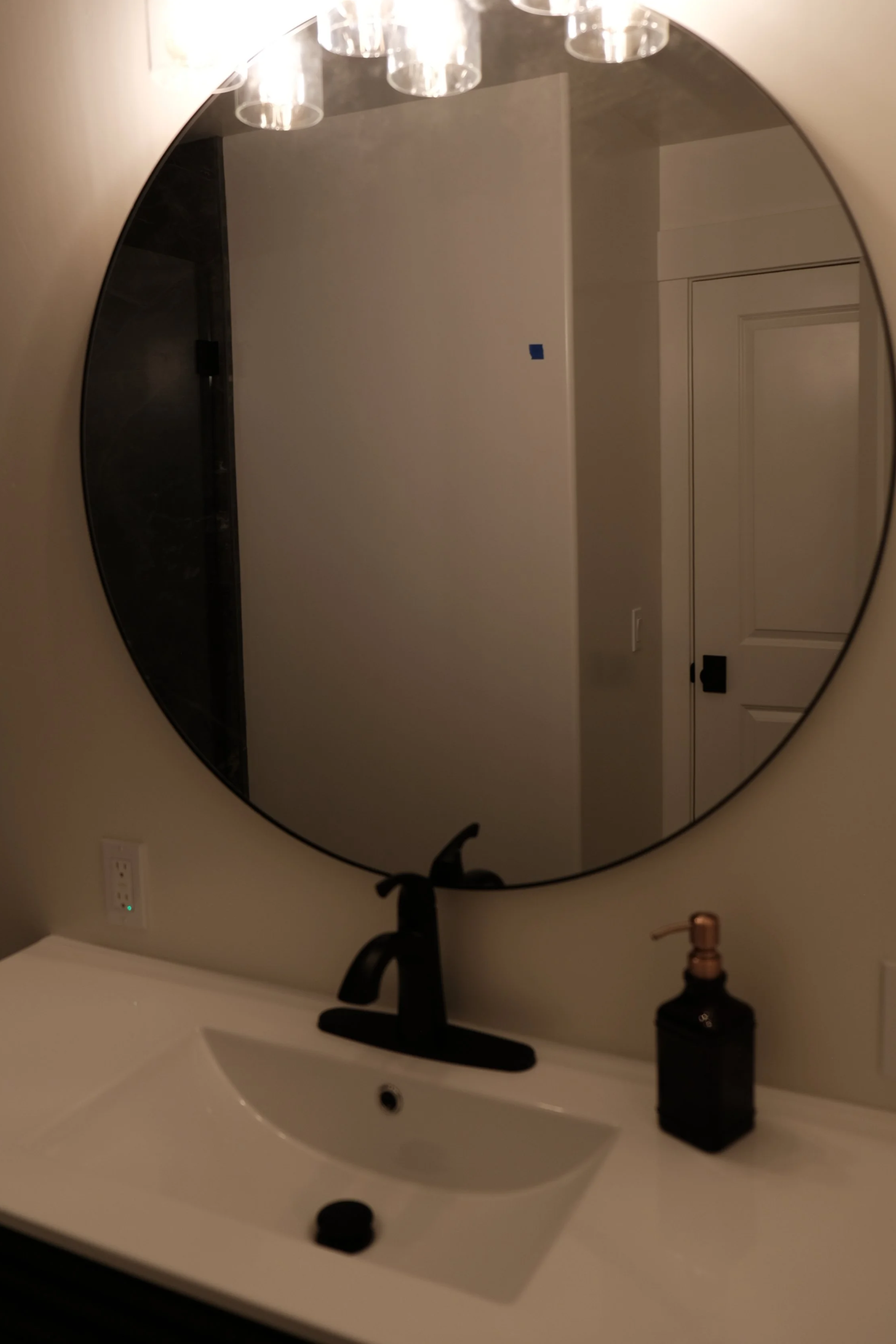 Bathroom mirror reflecting light fixture, door, and wall with a small blue square, above a white sink with black faucet and soap dispenser.
