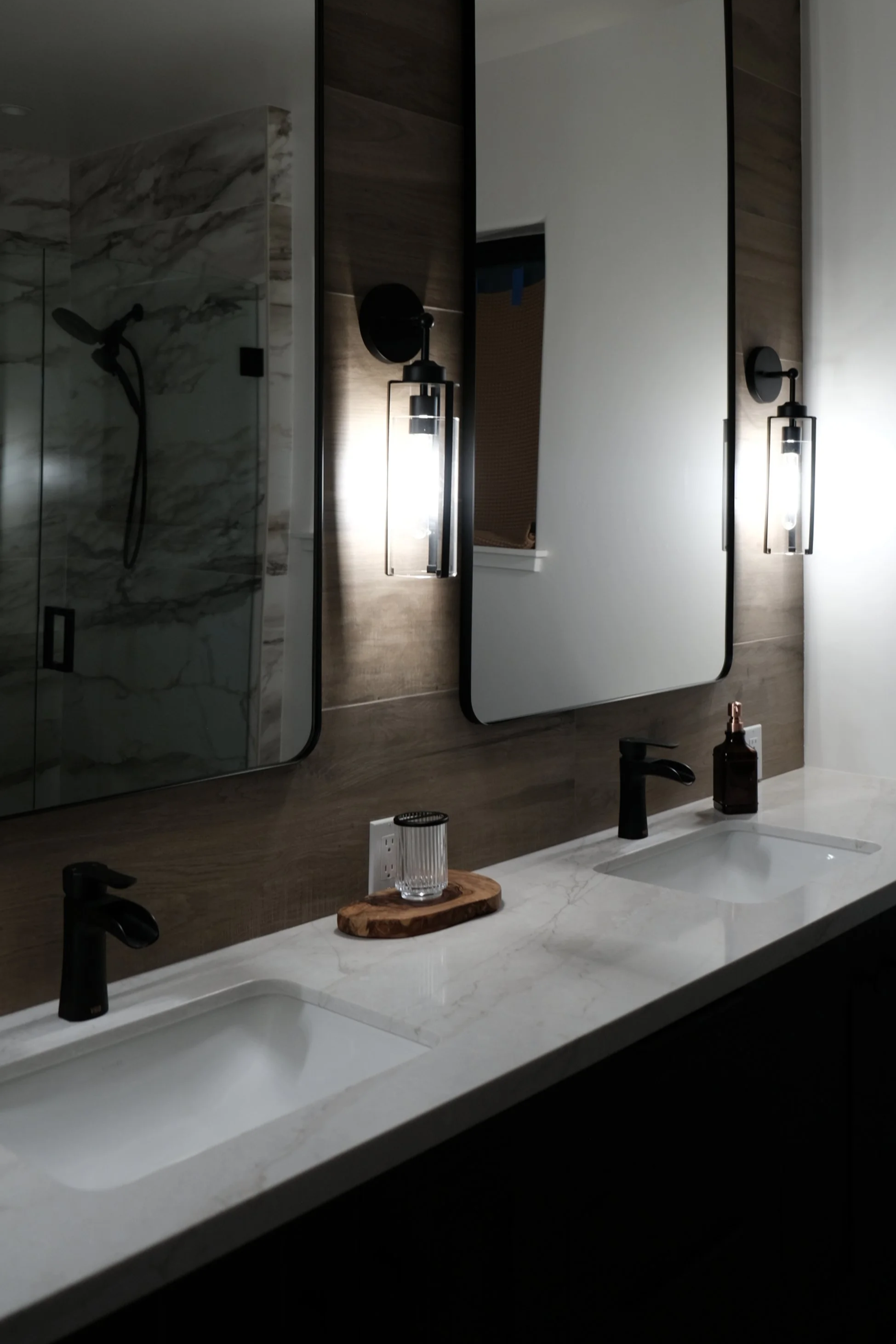 Modern bathroom with dual sinks, large mirrors, and wall-mounted light fixtures. There is a marble shower visible in the reflection of the left mirror.