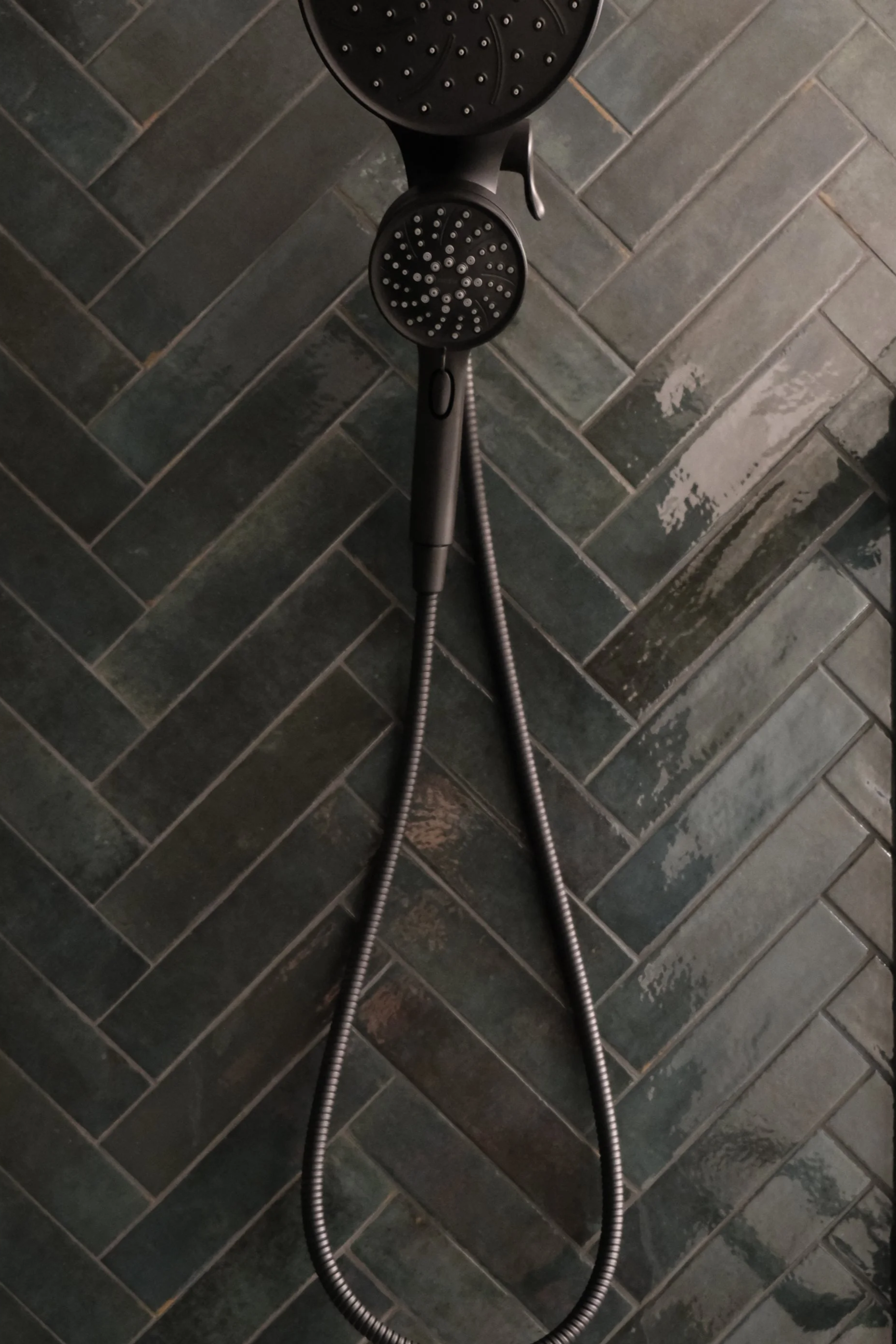 A black showerhead with a separate hand-held shower attached, mounted on a wall with dark gray, glossy, rectangular tiles arranged in a herringbone pattern.