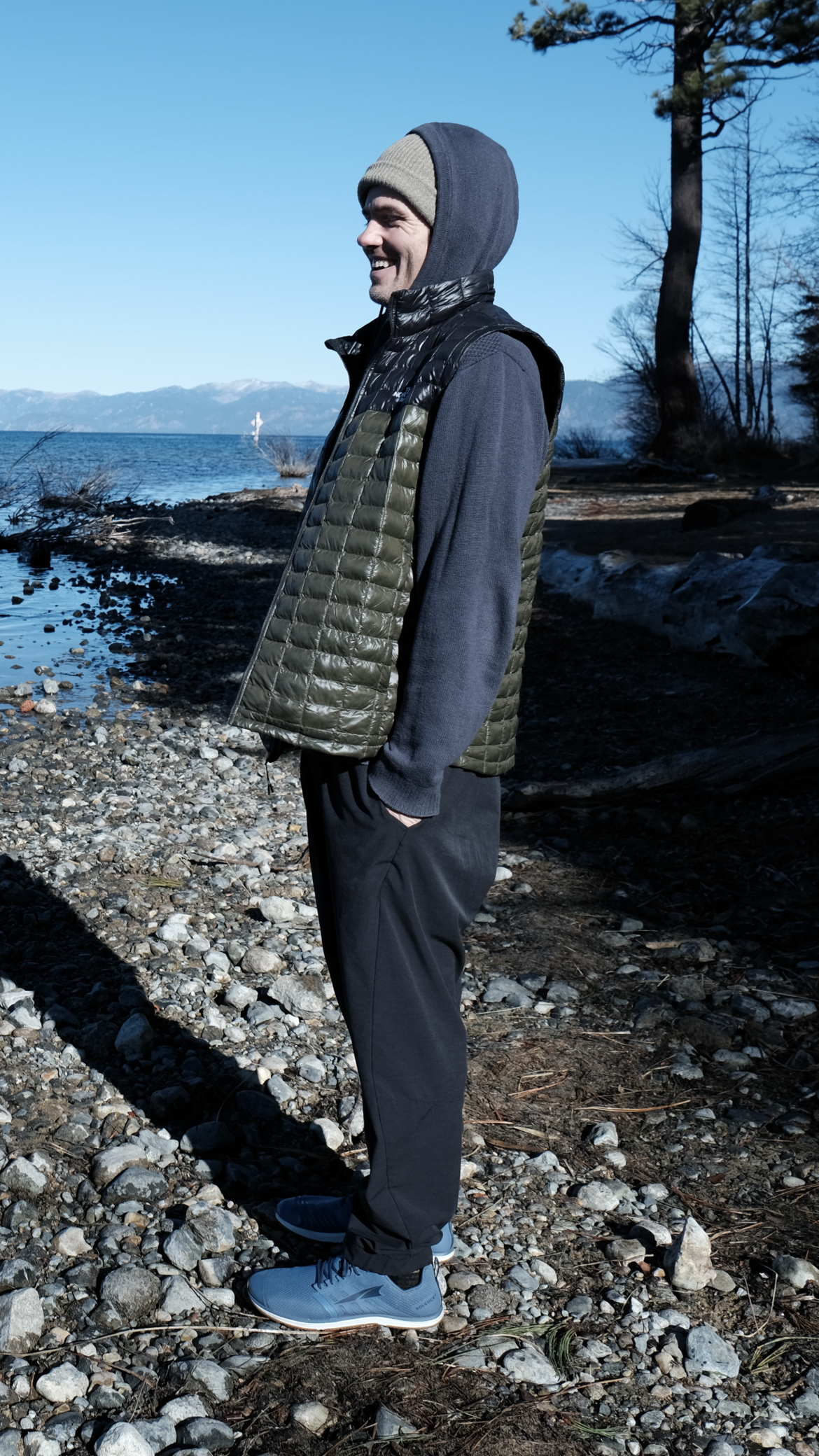 A man standing on a rocky lakeshore, wearing a gray beanie, black puffer vest, hoodie, black pants, and blue sneakers, smiling with a body of water and mountains in the background.