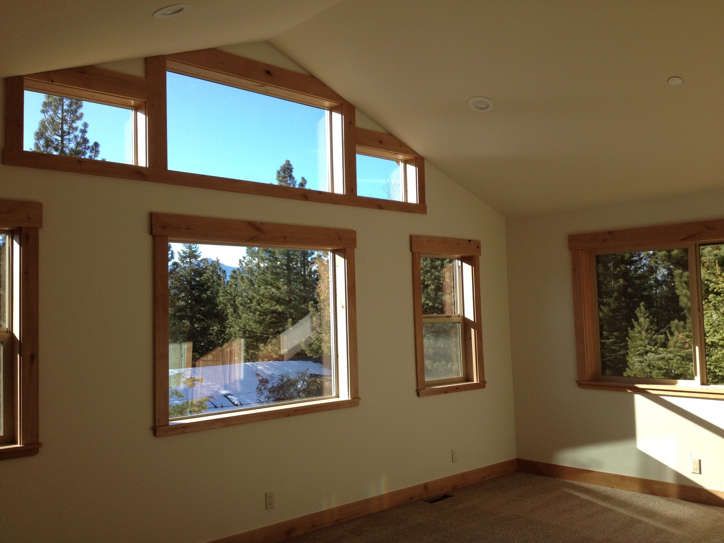 Empty room with large windows showing trees and blue sky outside, corner of the room with carpeted floor and wooden trim around the windows.