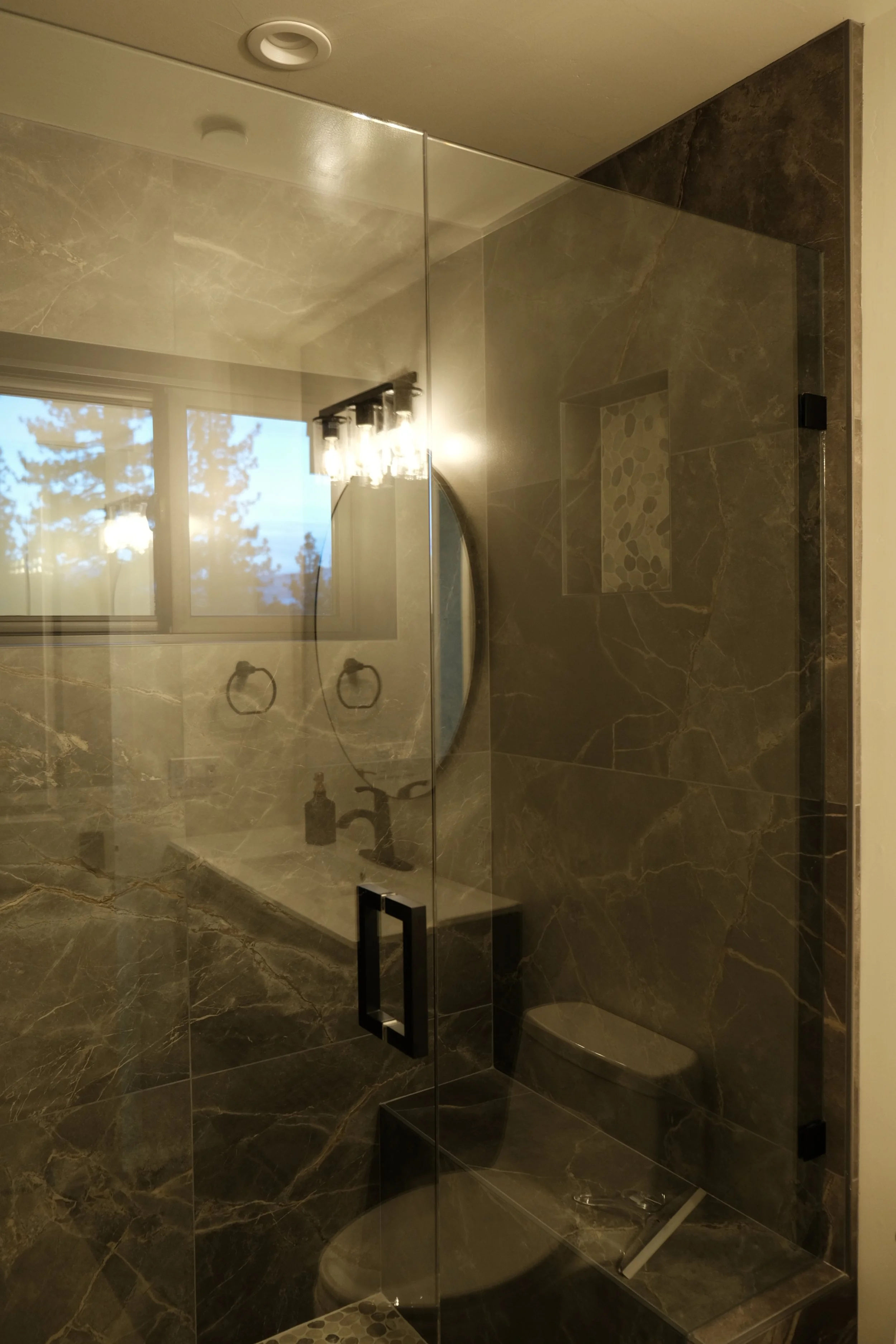 A bathroom with a glass shower enclosure, dark marble tiles, a window with trees outside, and a round mirror.