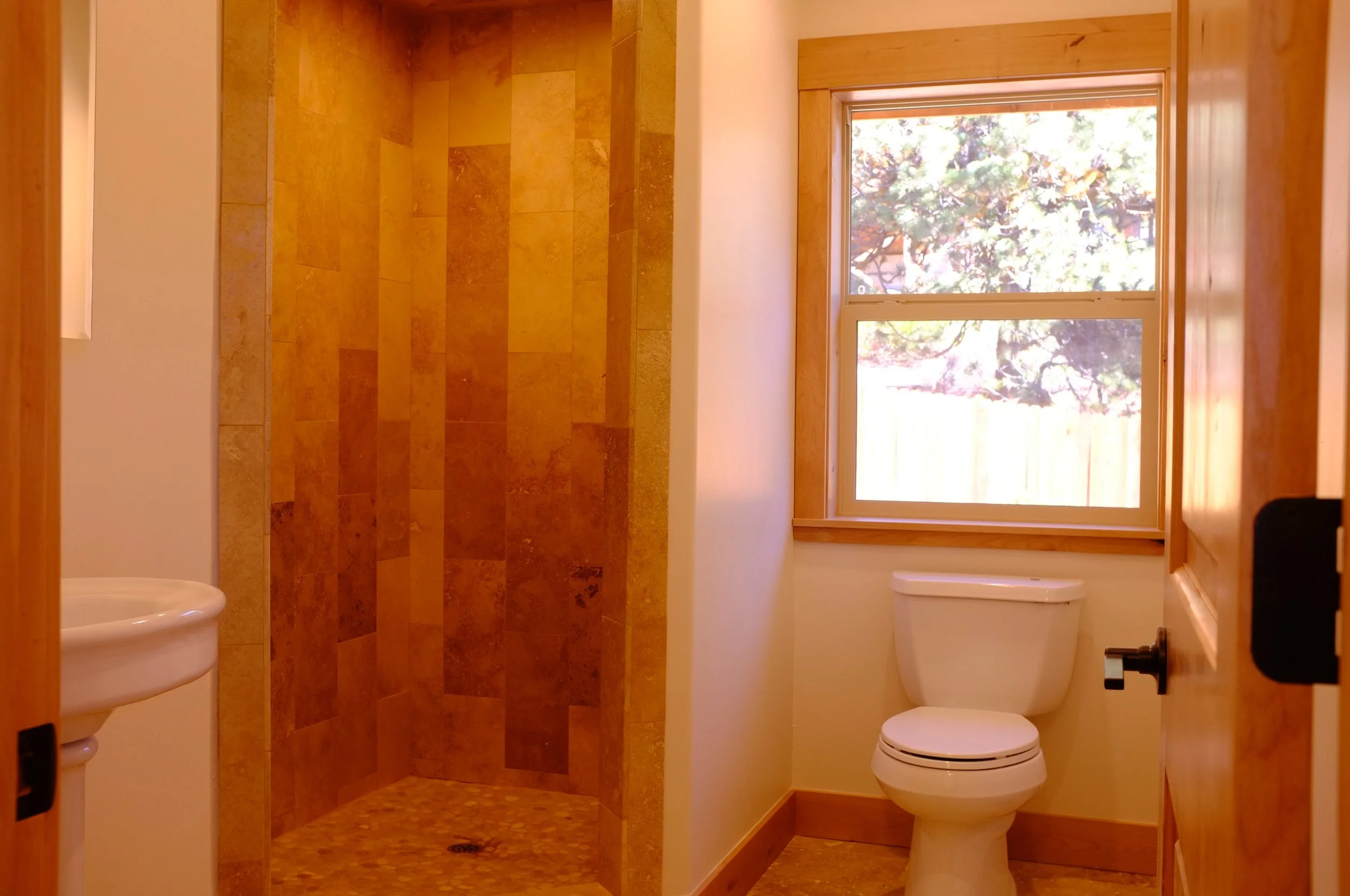 A small bathroom with a tan tile shower and a white toilet, window with a wooden frame, and a small portion of a white sink.
