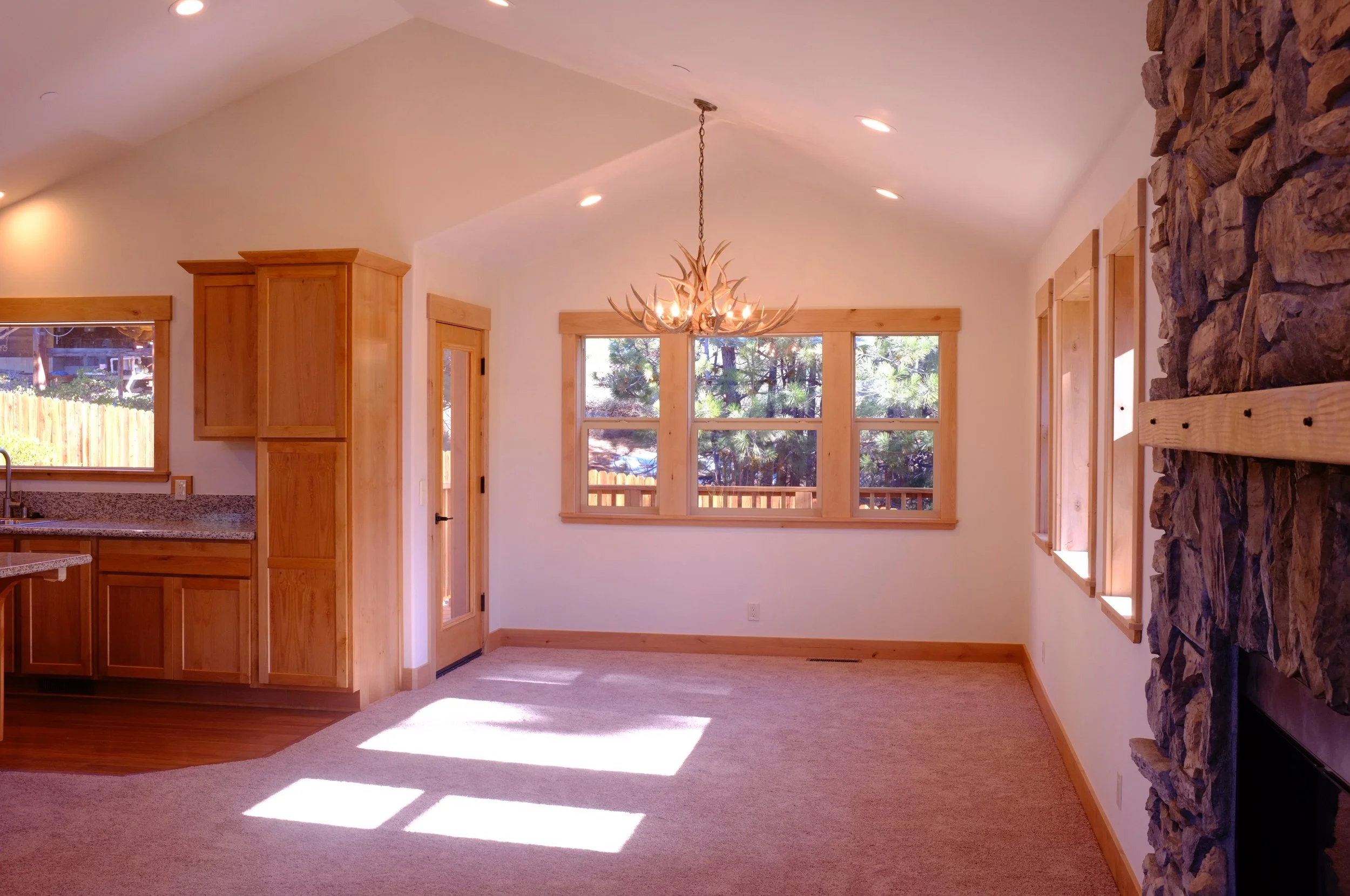 Empty living room with large windows, a stone fireplace on the right, wooden trim, and a chandelier hanging from the ceiling, illuminated by natural sunlight.
