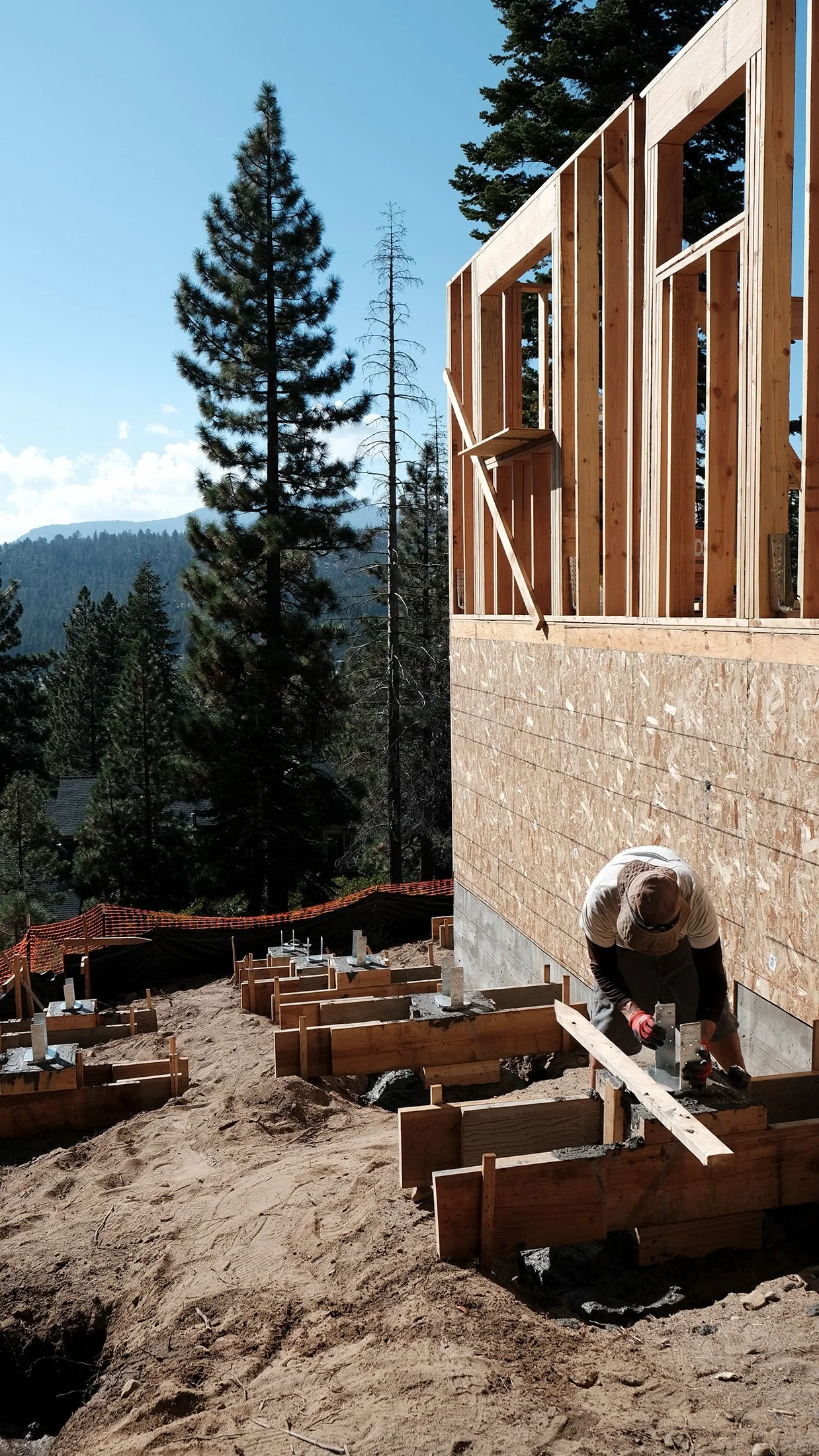 A construction worker is working on the foundation of a house on a hillside, with wooden framing being built on the upper part of the structure. Pine trees and mountains are visible in the background, indicating a forested, mountainous area.