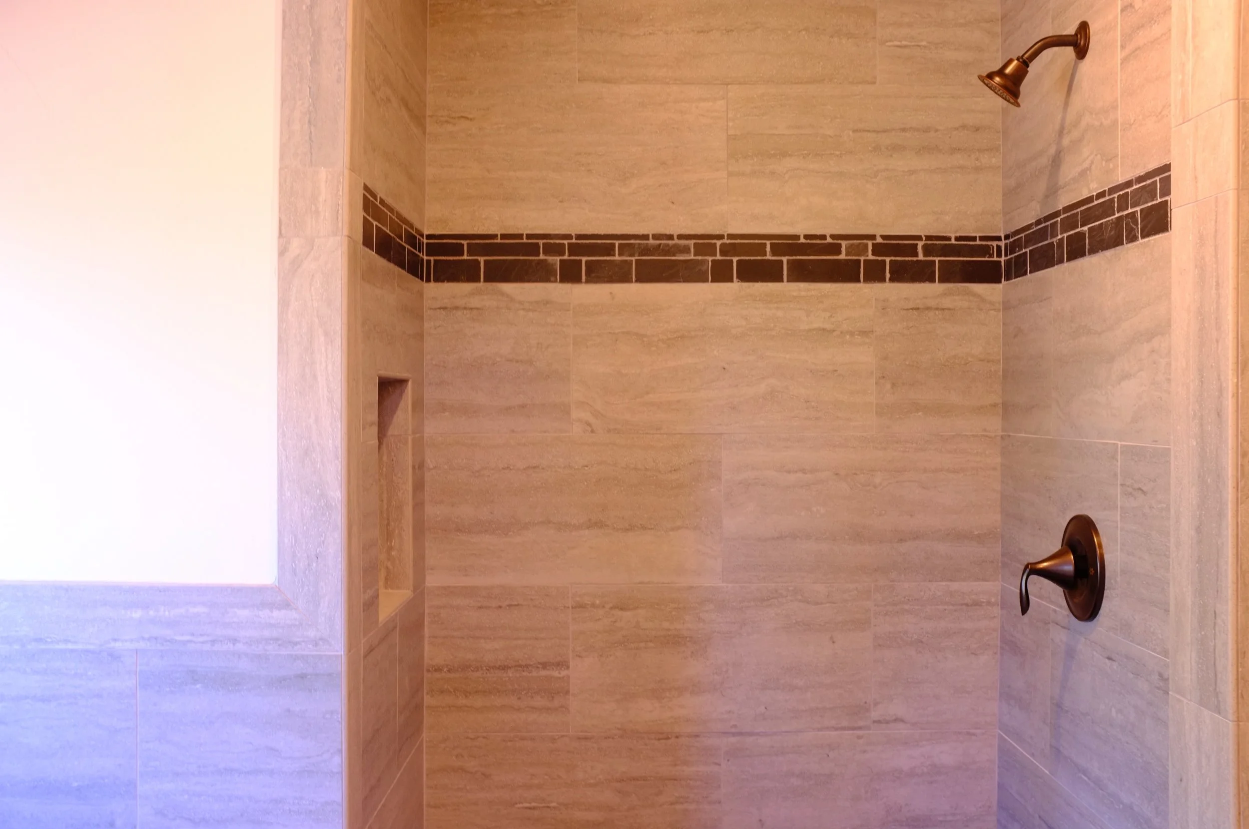 Empty shower with beige tiles, a decorative subway tile border, a small built-in shelf, a brass showerhead, and a brass faucet handle.