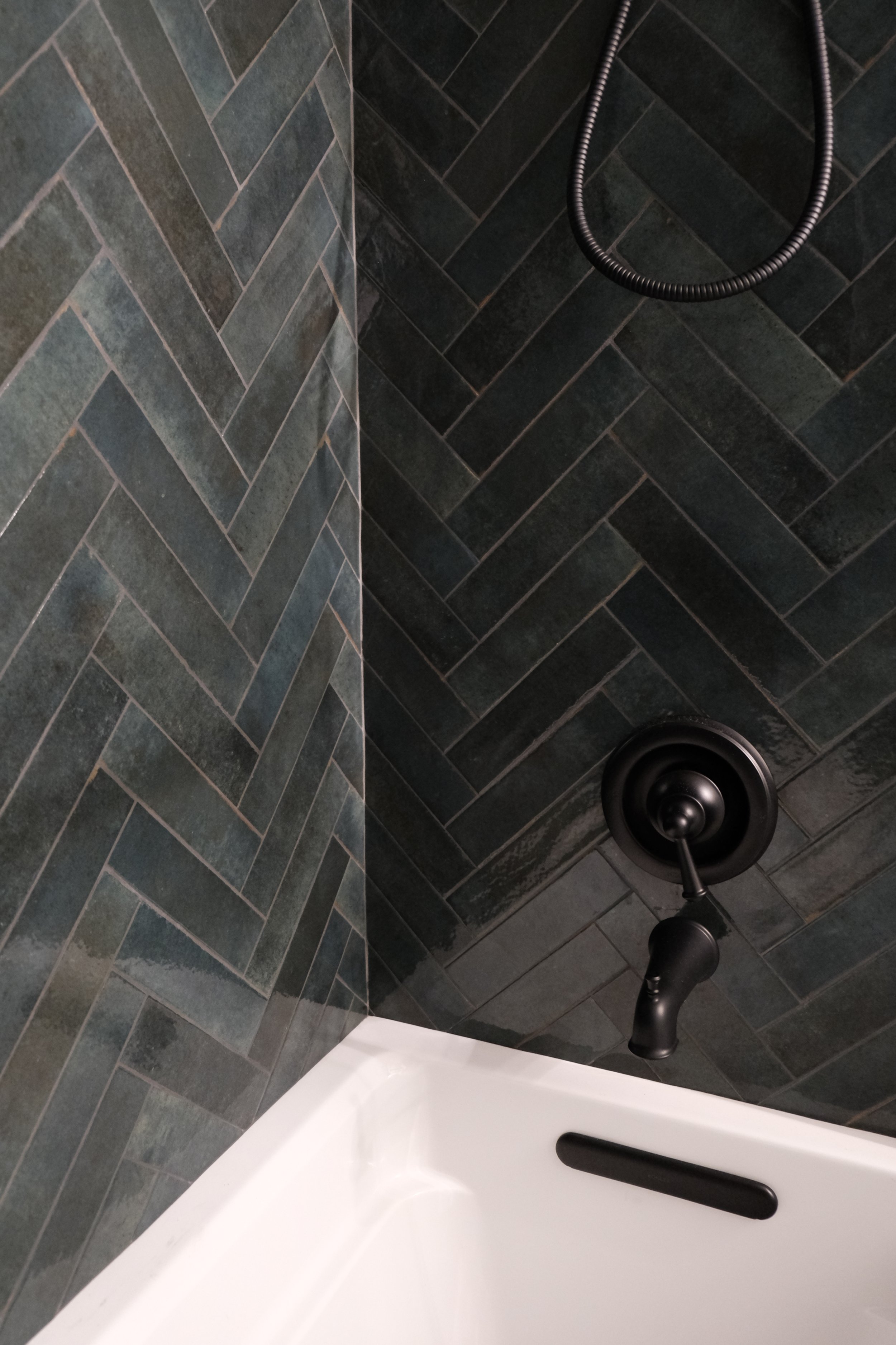Close-up of a shower area with dark herringbone patterned tiles, black shower fixture, and white bathtub edge.