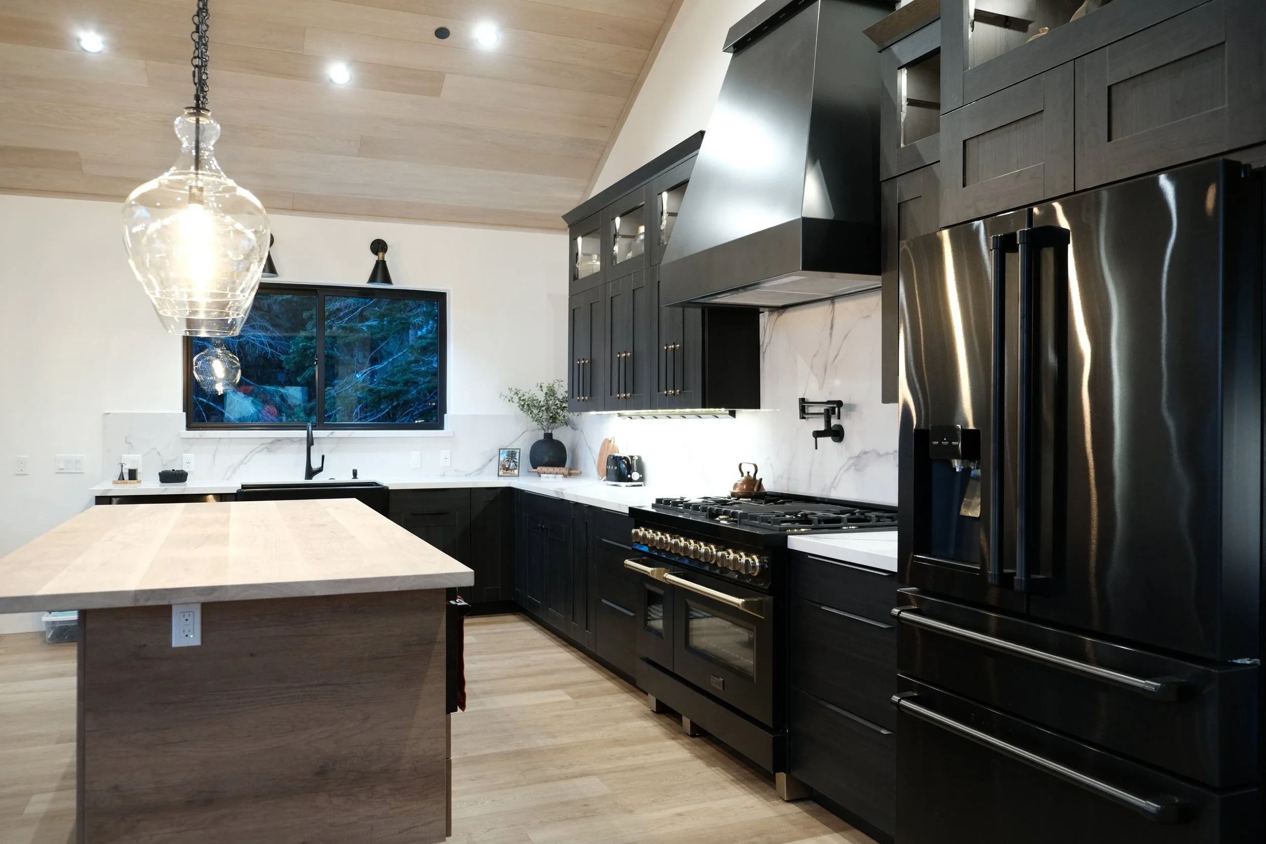 Modern kitchen with black cabinets, stainless steel appliances, a large island with a light wood countertop, a black refrigerator, black range oven, a window overlooking trees, and pendant lighting.