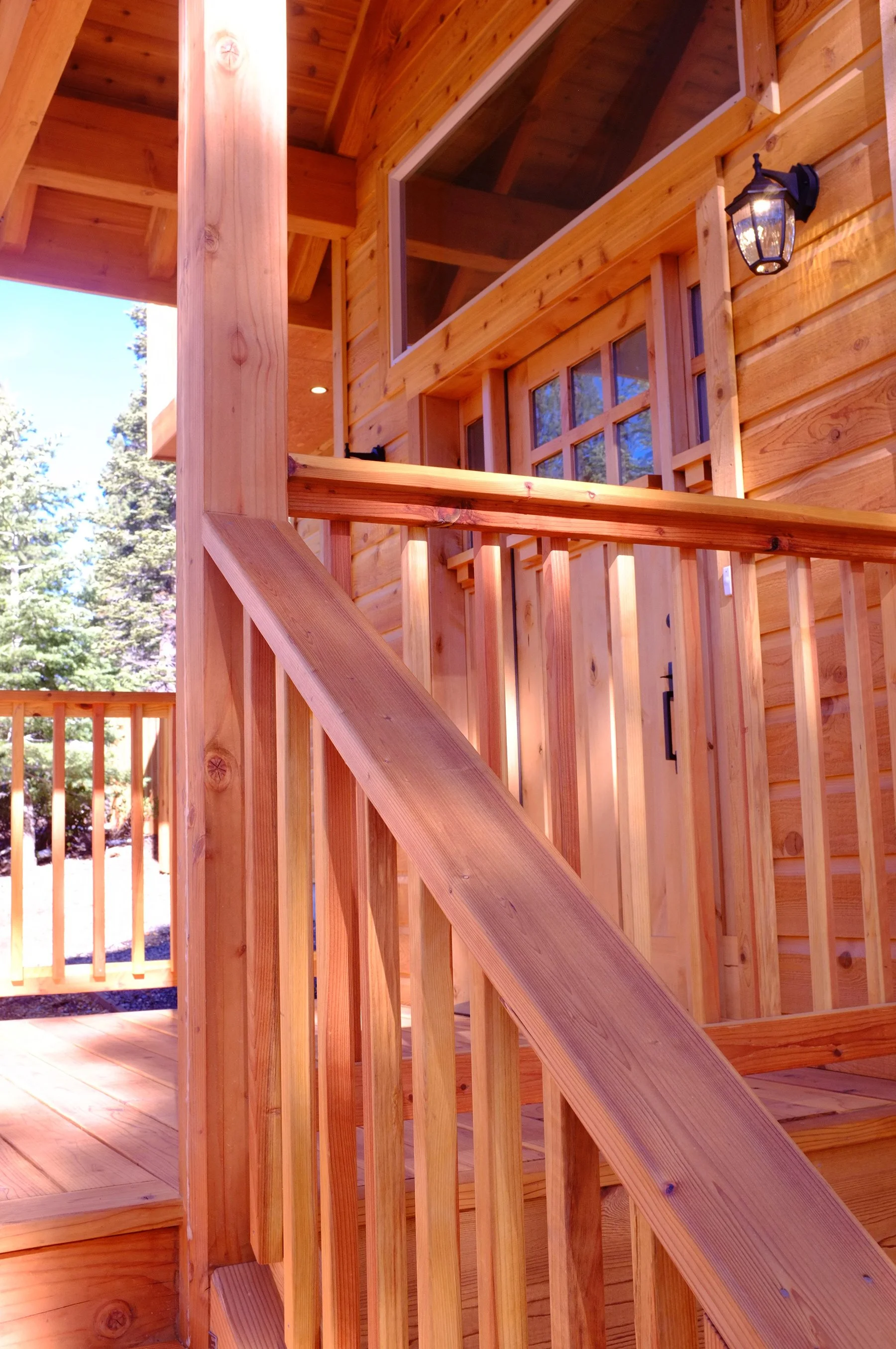 Close-up of a wooden balcony with a railing and steps, showing the exterior of a log cabin with large windows and a lantern-style outdoor light fixture, surrounded by trees.