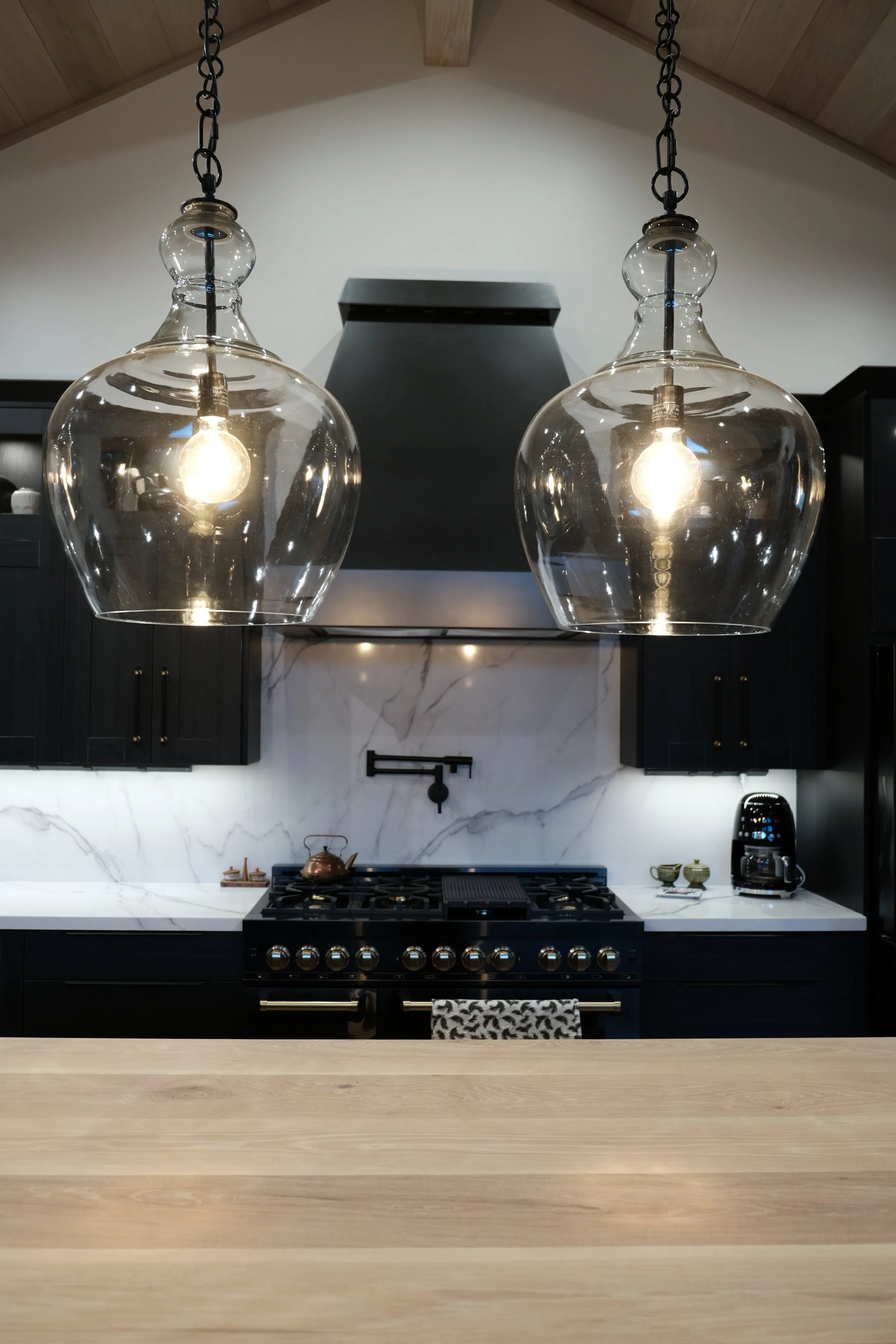 Two glass pendant lights hanging over a modern kitchen with black cabinets, white marble backsplash, and a black stove with a vent hood.